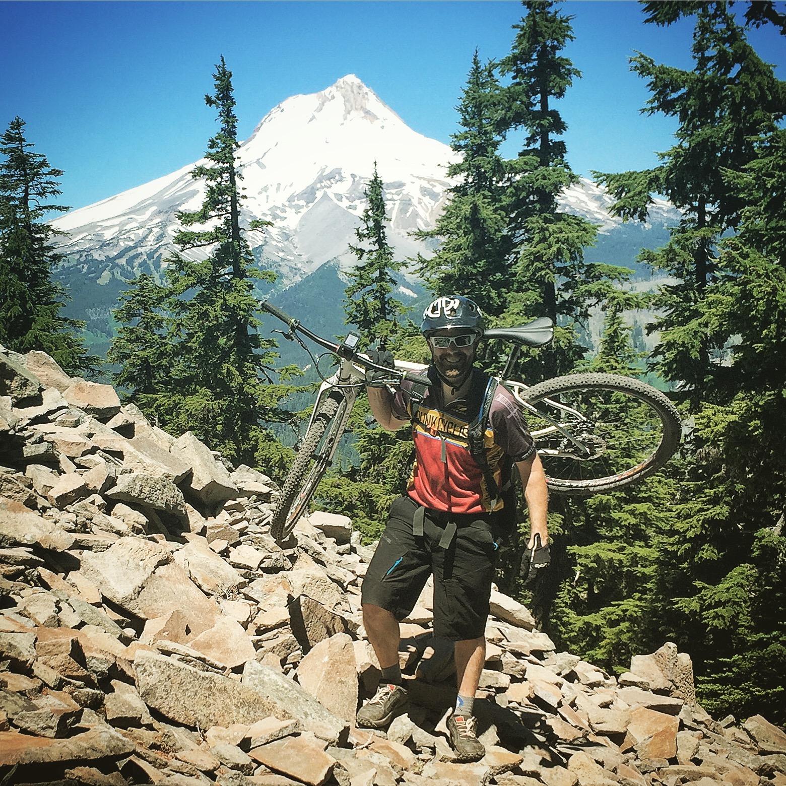 Niner S.I.R. 9: A mountain biker carrying his bike over rocky terrain with a forest of pine trees in the background and a snow-capped mountain peak visible in the distance.