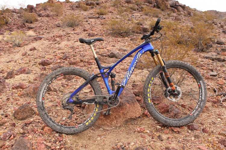 A blue mountain bike resting on rocky terrain, surrounded by sparse vegetation and arid landscape. The bike features thick tires and a suspension fork, indicating it is designed for off-road use.