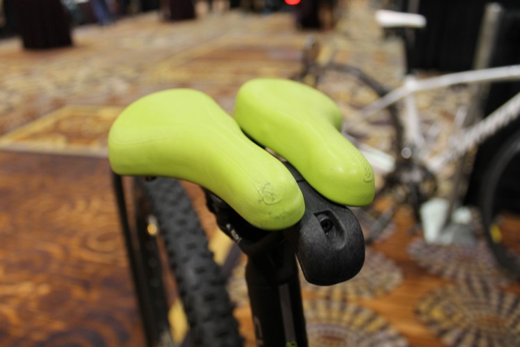 Image of a bicycle seat featuring a unique design with two green, contoured saddle pads mounted on a black seat post. The background shows a blurred indoor setting, indicating a possible exhibition or event.
