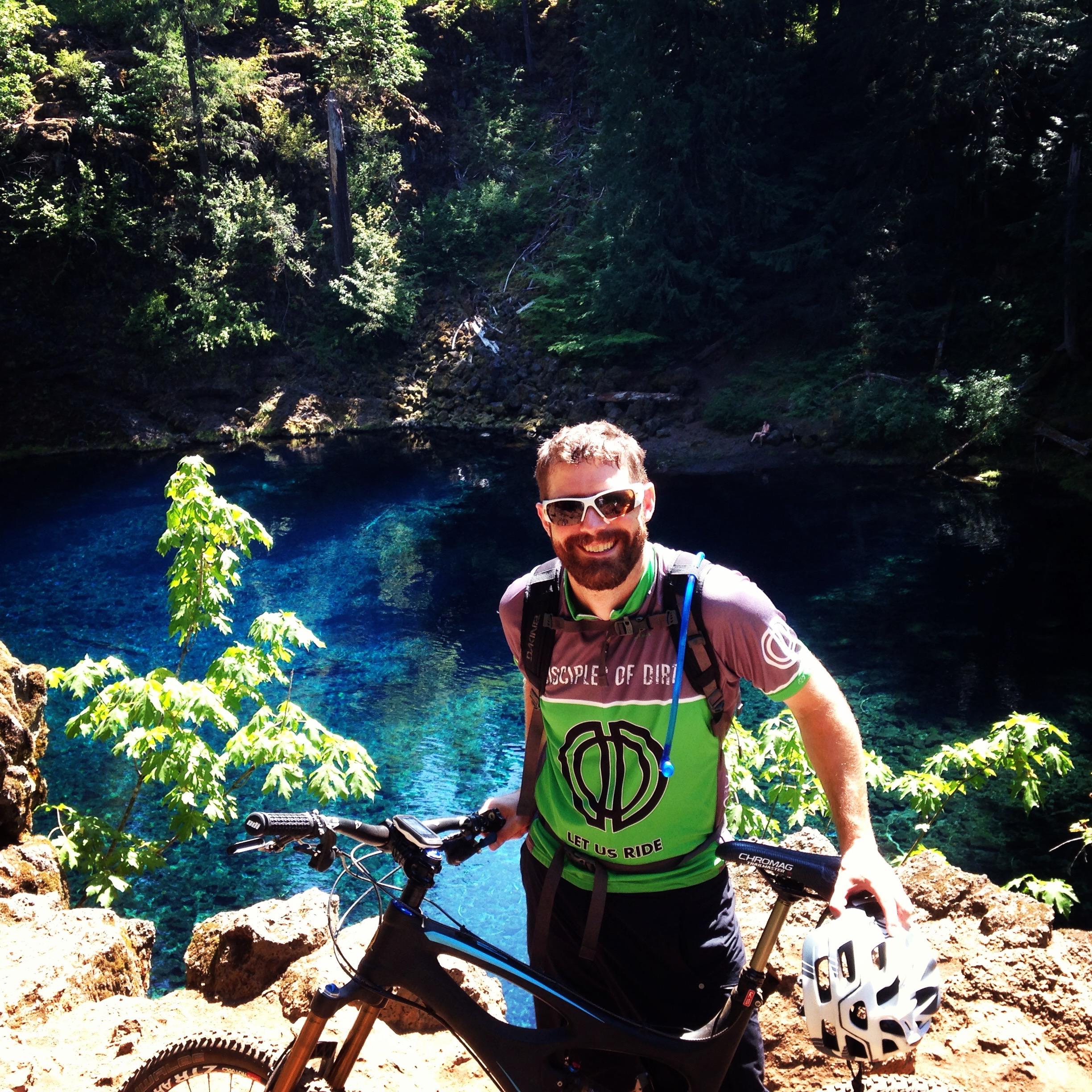 A man stands next to his mountain bike, smiling while wearing a green and grey athletic shirt, helmet in hand. Behind him is a clear blue body of water surrounded by lush greenery and rocky terrain. Sunlight filters through the trees, creating a vibrant outdoor scene.