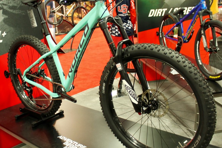 A turquoise mountain bike displayed on a black platform at an exhibition, featuring thick tires and a prominent front suspension fork. The background includes blurred images of other bikes and promotional materials.