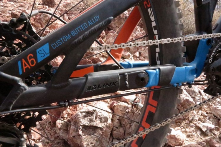 Close-up view of a mountain bike frame featuring a custom-butted alloy construction, with visible components such as the chain, rear derailleur, and tire. The image highlights a bright blue link piece and orange accents on the frame against a rocky terrain background.