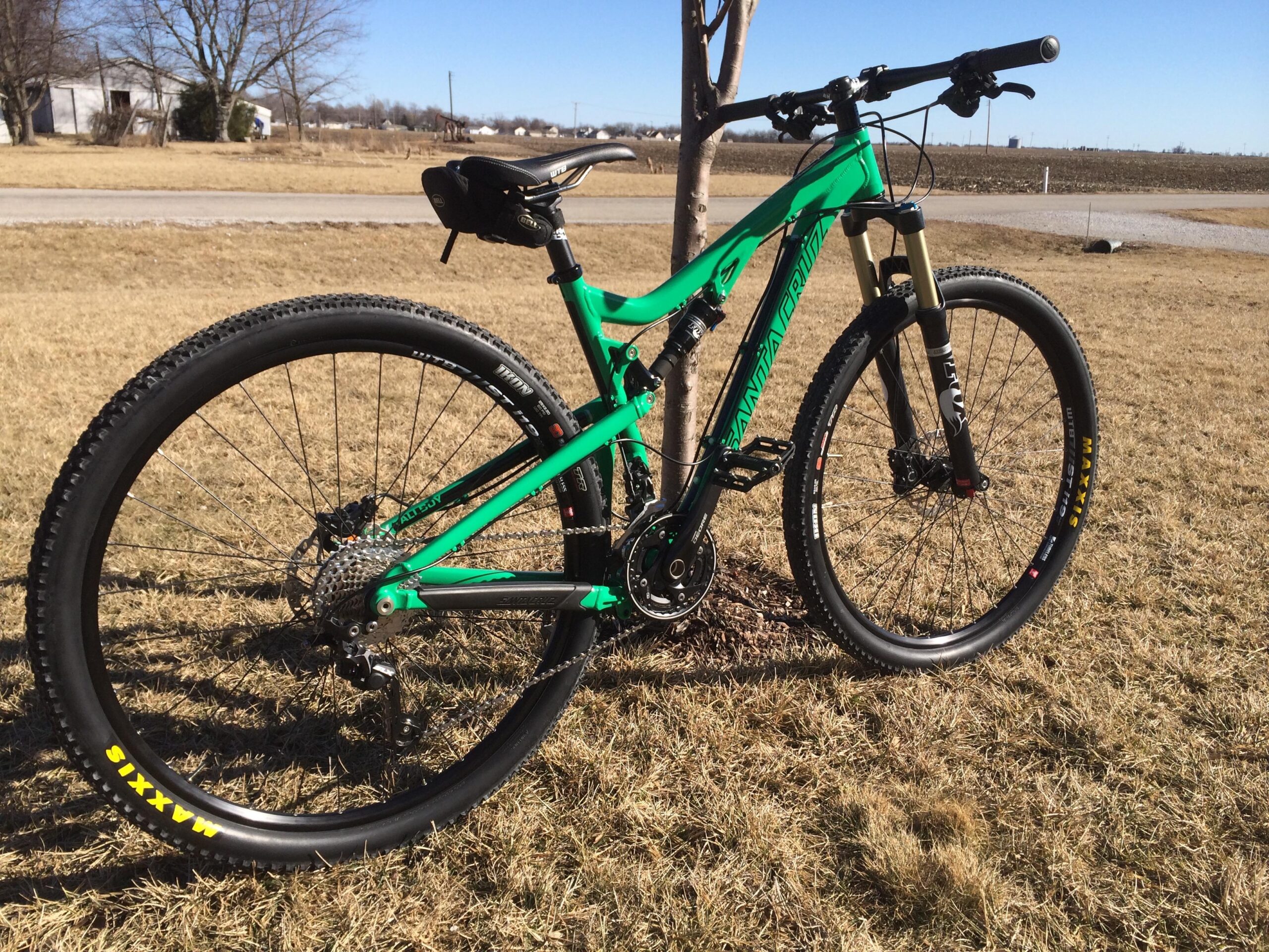 Santa Cruz Tallboy: A brightly colored green mountain bike stands next to a small tree on a grassy area, with a clear blue sky in the background. The bike features thick tires and a sturdy frame, showcasing its off-road capabilities. In the distance, there are hints of farm buildings and open fields.