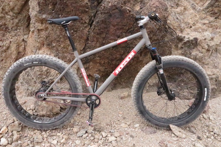 A gray fat bike leaning against a rocky surface, featuring large tires for improved traction. The bike has a simple, robust design, with a black seat and handlebar, and a chain visible near the pedals. The background consists of rugged terrain with loose gravel and rocks.