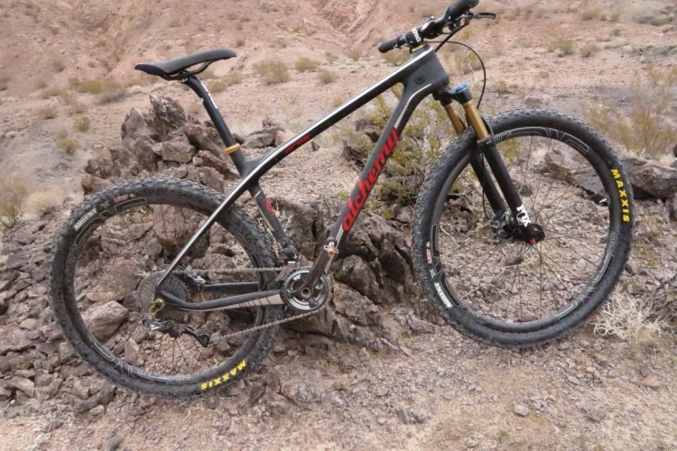 A mountain bike leaning against a rocky outcrop in a rugged, arid landscape. The bike features a sleek design with black and red accents, showcasing high-quality components including wide tires and a sturdy frame. The background consists of rocky terrain and sparse vegetation, typical of a desert environment.