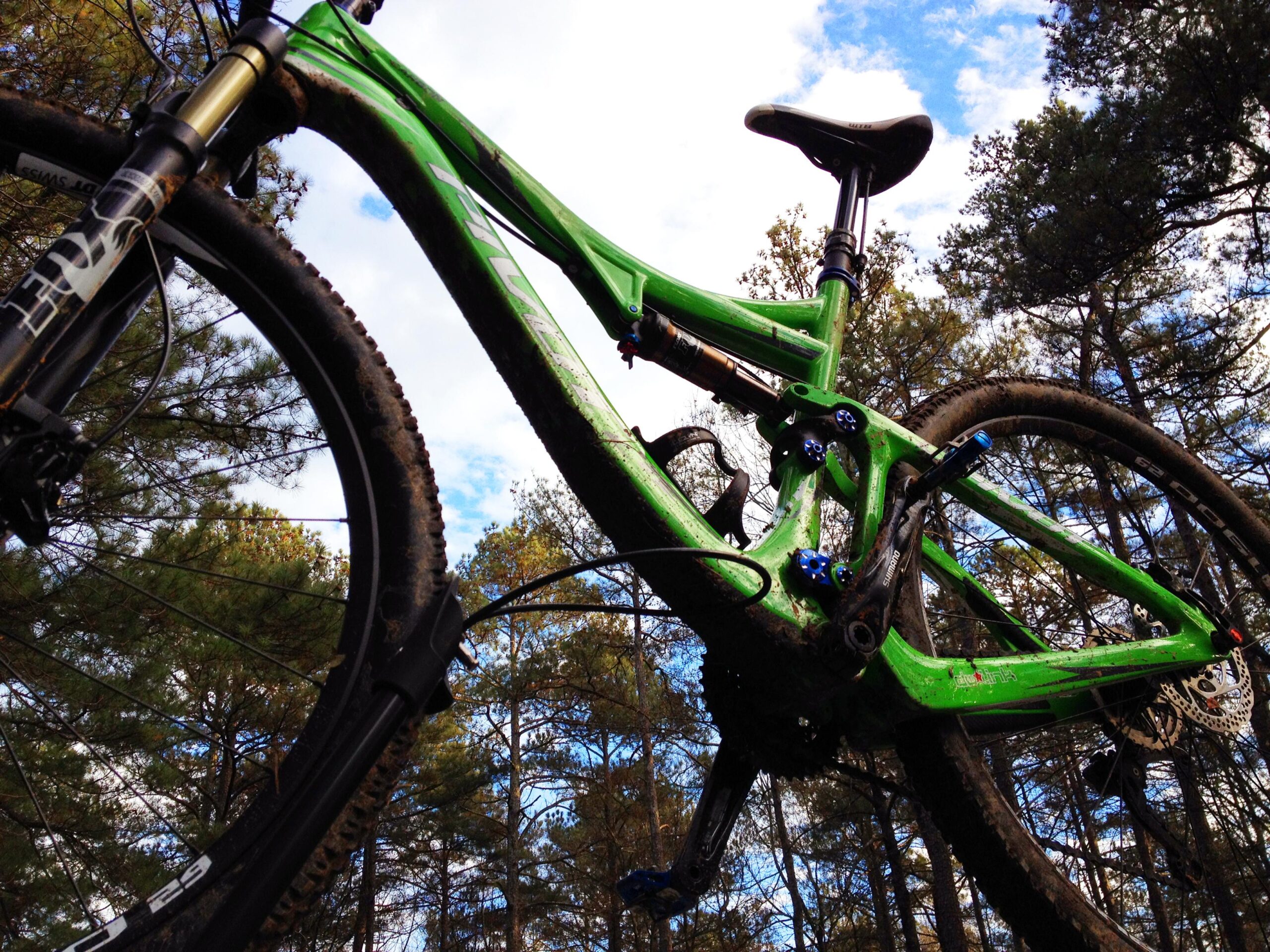 Pivot Mach 429 Carbon: A close-up view of a green mountain bike with muddy tires, captured from a low angle against a backdrop of tall trees and a cloudy sky. The bike appears to be in a natural outdoor setting, showcasing its rugged design and readiness for trail riding.