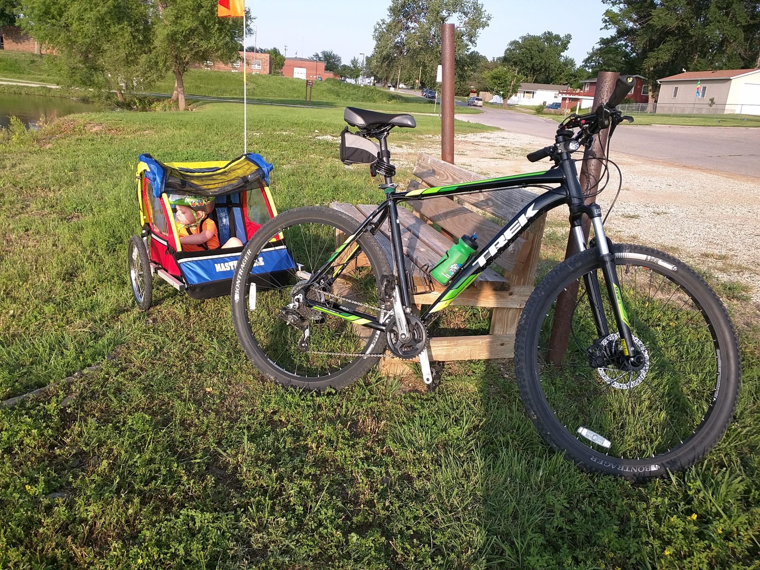 Trek Marlin 6: A Trek mountain bike is parked next to a wooden bench, with a child sitting comfortably in a colorful bike trailer attached to the bike. The scene is set in a grassy area near a small pond, with a few trees and buildings in the background under clear blue skies.