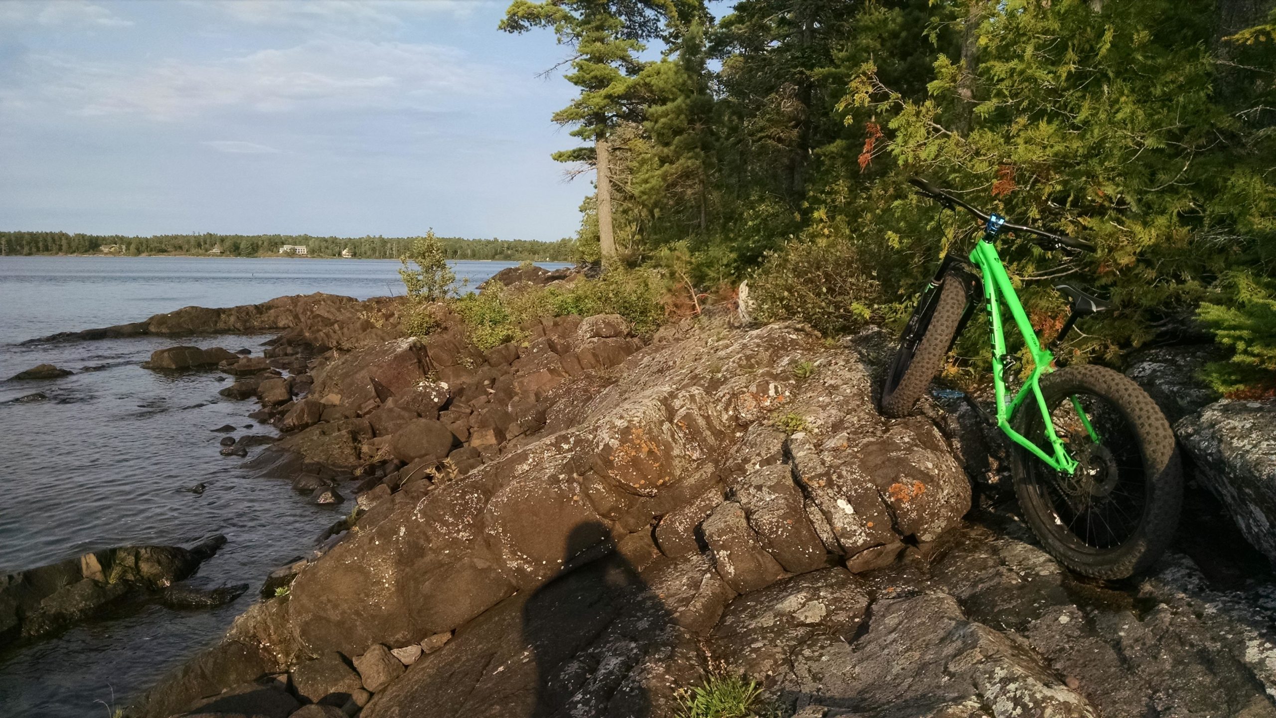 A bright green fat bike rests on rocky terrain near a calm lake, surrounded by trees and under a blue sky. The shoreline is visible, with gentle waves lapping against the rocks in the foreground. Copper Harbor Trails mountain bike trail.