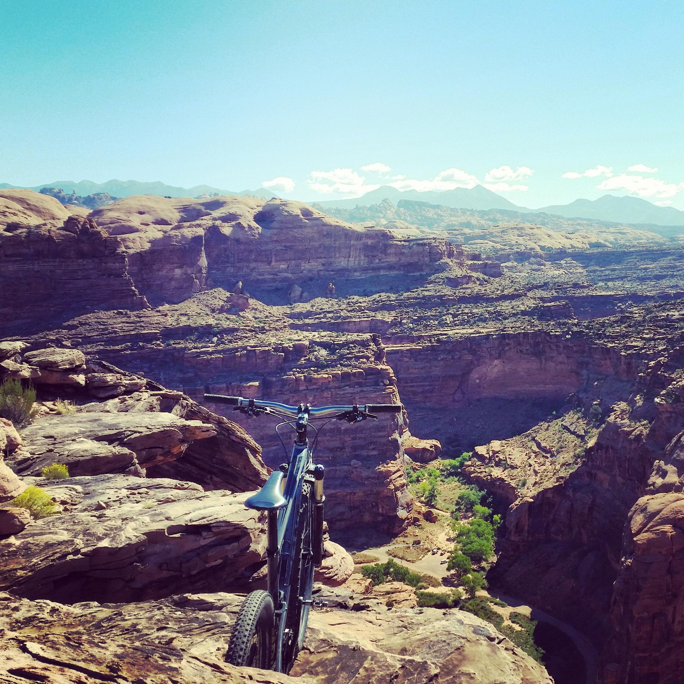 A mountain bike perched on the edge of a rocky cliff, overlooking a deep canyon with layered rock formations. The scene features a clear blue sky and distant mountains in the background, creating a sense of adventure and breathtaking natural beauty. Captain Ahab mountain bike trail.