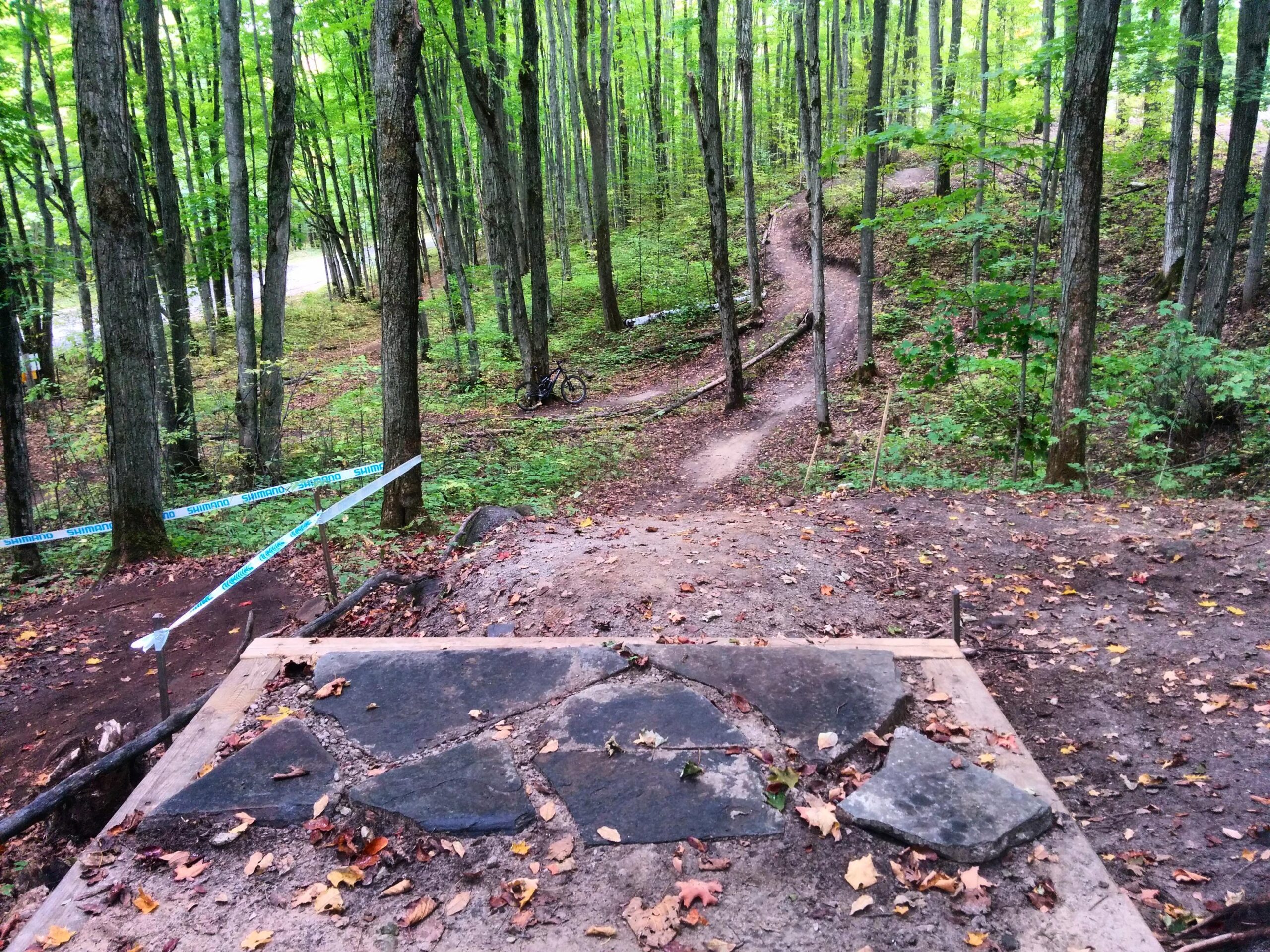 Mountain biking trail in a forest setting, featuring a stone platform at the forefront, with a winding dirt path leading through trees in the background. A bike is parked nearby, and fallen leaves are scattered on the ground. Hardwood Ski and Bike mountain bike trail.