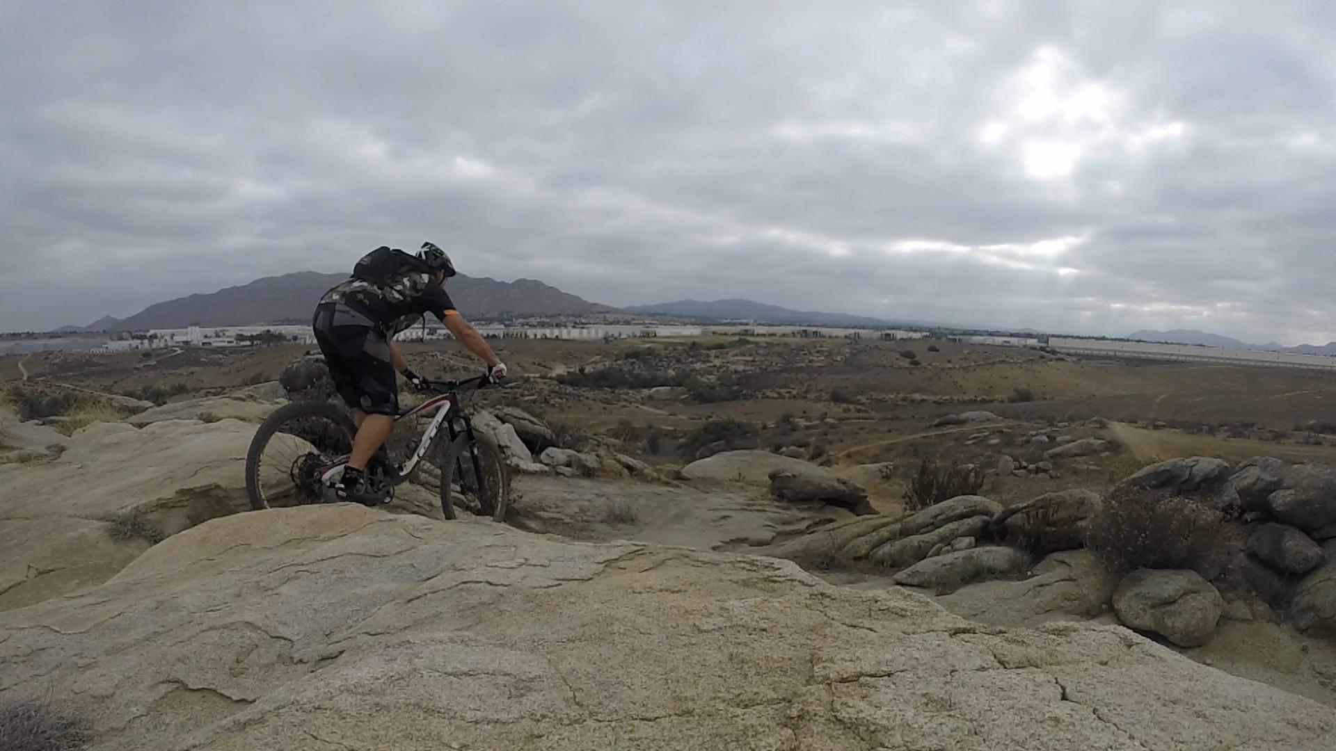 A mountain biker navigating rocky terrain with a backdrop of rolling hills and cloudy skies. The rider is focused on the path ahead, showcasing a dynamic outdoor activity in a natural setting. Sycamore Canyon Park mountain bike trail.