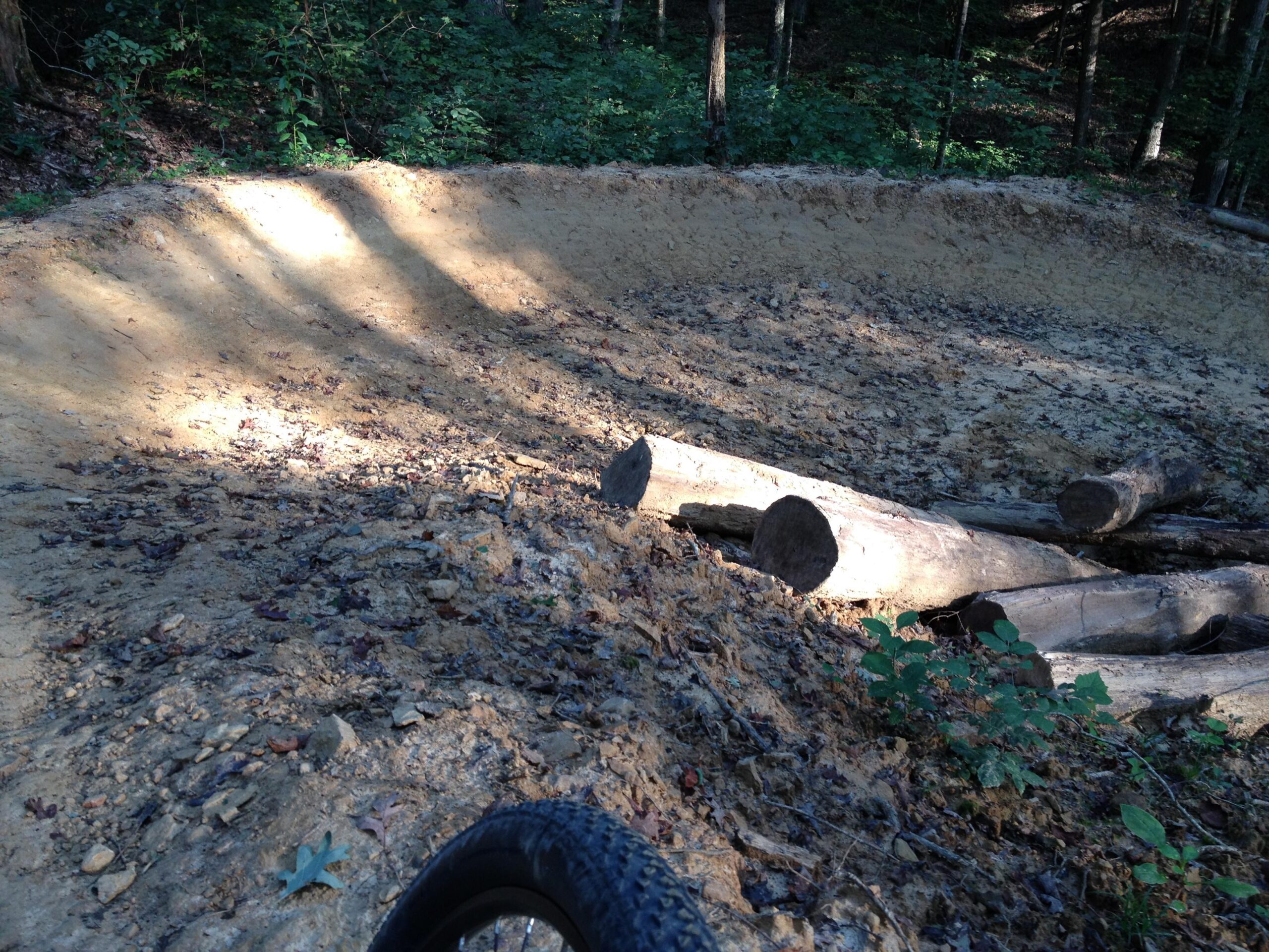 A dirt bike trail with a curved shape, surrounded by trees and foliage. Logs are placed along the edge of the trail, and sunlight casts shadows on the packed dirt surface, indicating a well-used path. A portion of a bicycle tire is visible in the foreground. O'bannon Woods mountain bike trail.