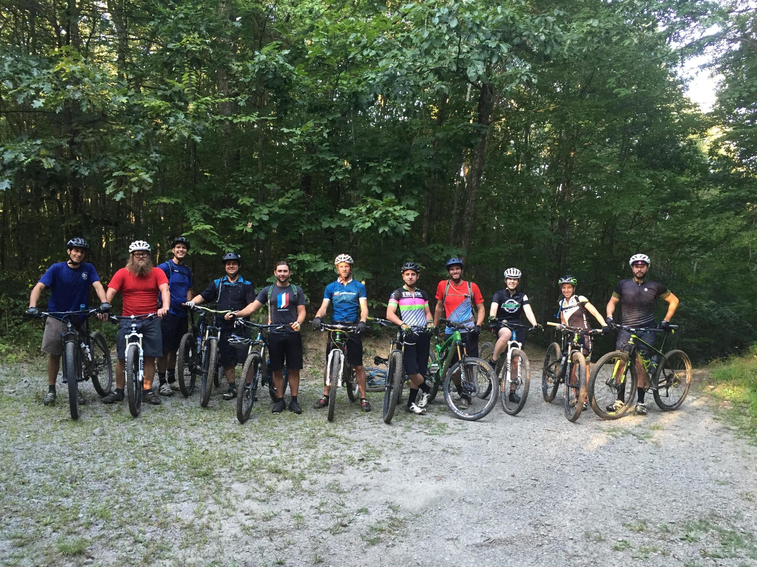 A group of eleven mountain bikers poses together with their bikes on a gravel path surrounded by lush green trees. They are dressed in casual cycling attire, wearing helmets, and smiling for the camera. The atmosphere suggests a friendly camaraderie among outdoor enthusiasts ready for an adventure.