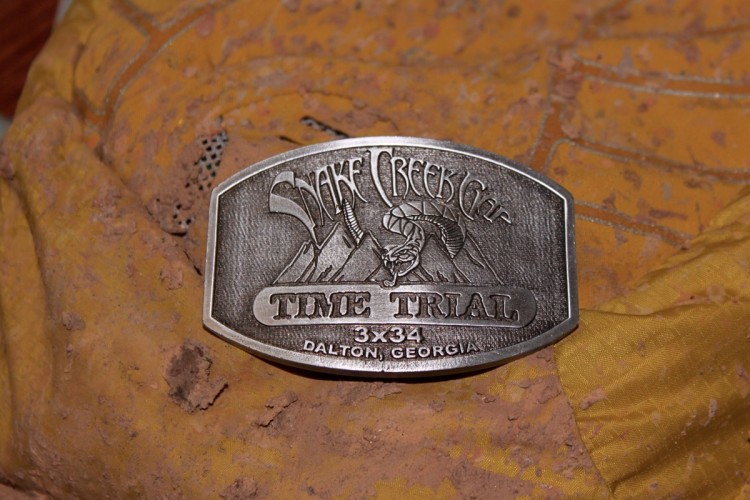 A close-up view of a silver medal for the "Snake Creek Gap Time Trial," featuring intricate engravings of mountains and a snake, placed on a textured, yellow fabric background covered in dirt. The bottom of the medal indicates "3x34" and "Dalton, Georgia."