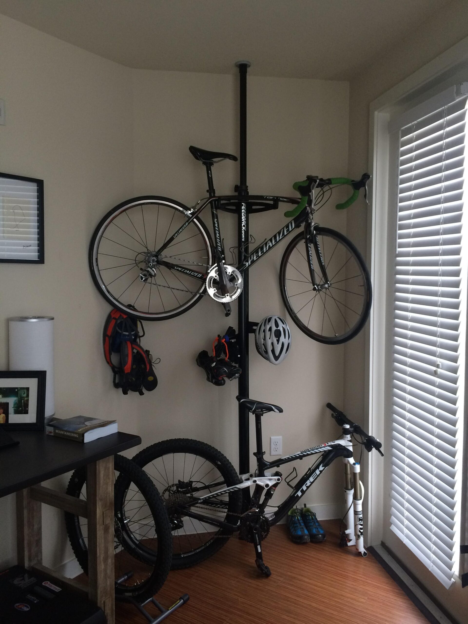 Trek Remedy 8 27.5/650b: A corner of a room featuring two bicycles securely stored. A road bike is mounted on the wall with a vertical storage pole, while a mountain bike is leaned against a table. Nearby, there are helmets and accessories hanging from the wall. Natural light filters in through a window with blinds, illuminating the space.