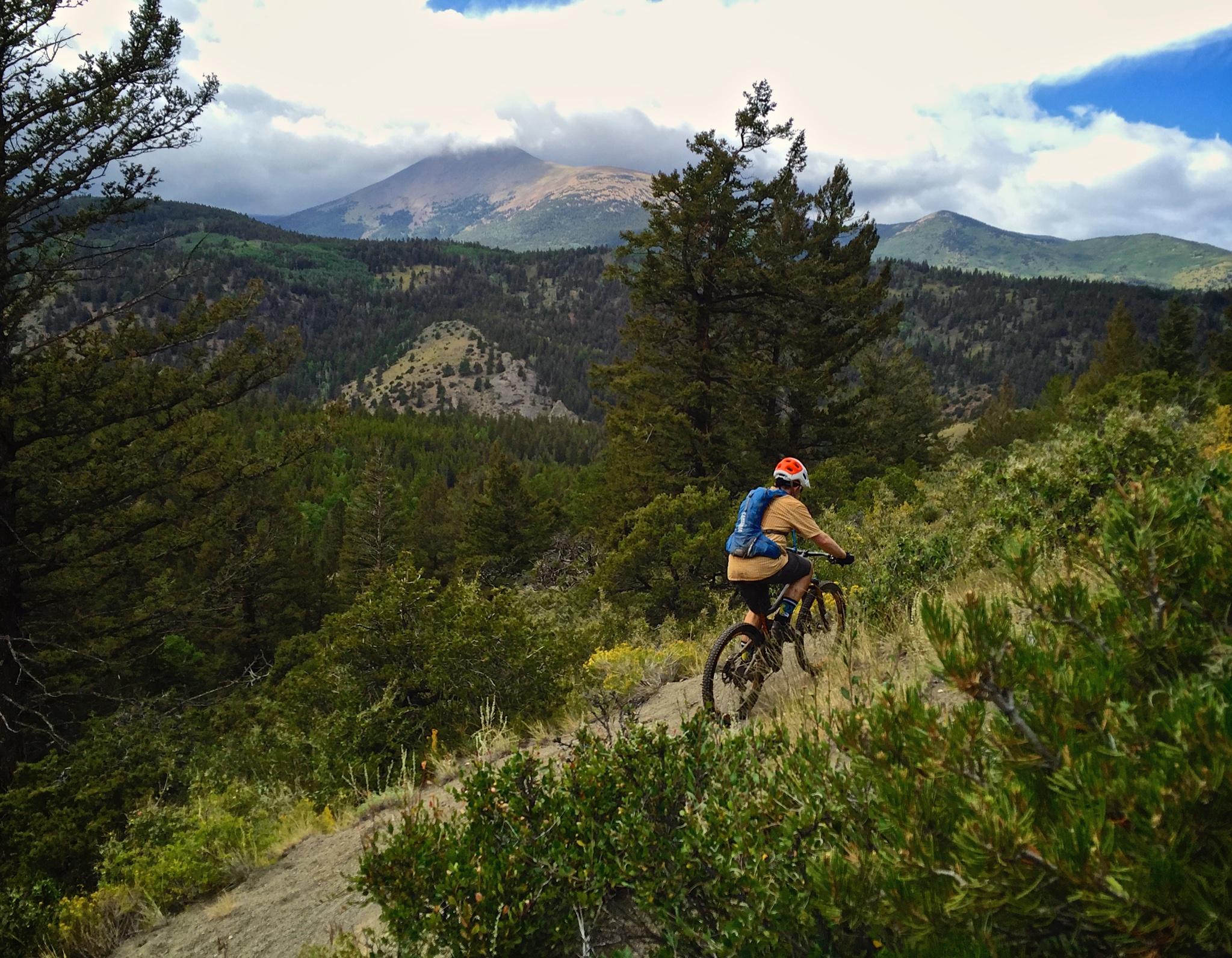 A mountain biker riding along a rugged trail surrounded by lush green trees and distant mountains under a partly cloudy sky. Monarch Crest Trail mountain bike trail.