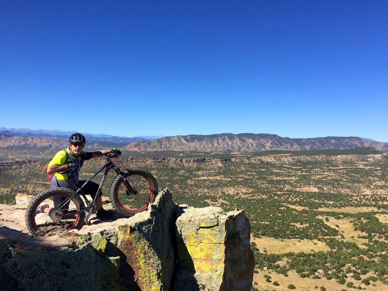 A mountain biker poses with their bike on a rocky ledge, overlooking a vast landscape of rolling hills and mountains under a clear blue sky. The scene captures the spirit of adventure in an outdoor setting. Oil Well Flats mountain bike trail.