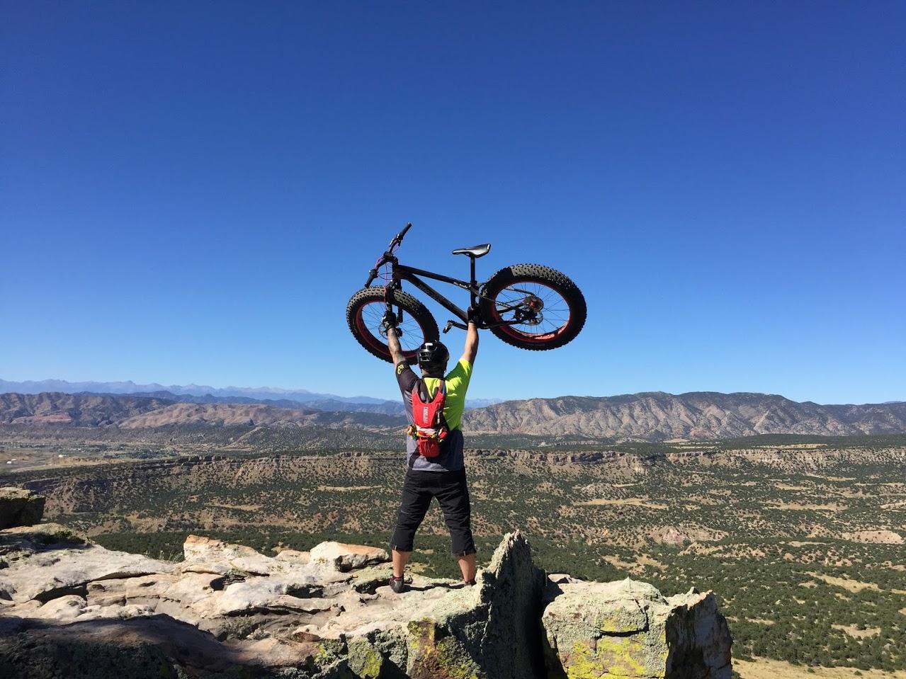 A mountain biker standing triumphantly on a rocky outcrop, holding a fat bike overhead. The expansive landscape features rolling hills and mountains under a clear blue sky. Oil Well Flats mountain bike trail.