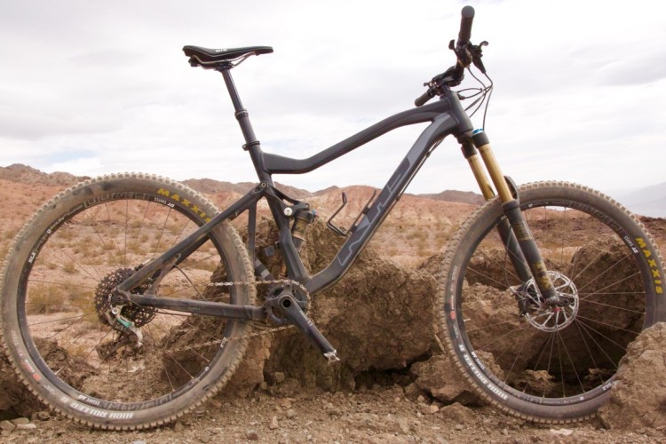A black mountain bike is positioned against a rocky outcrop in a desert landscape, featuring rugged terrain, dry bushes, and distant hills. The bike showcases front and rear suspension, wide tires, and a streamlined frame designed for off-road riding. The sky is partly cloudy, enhancing the outdoor setting.
