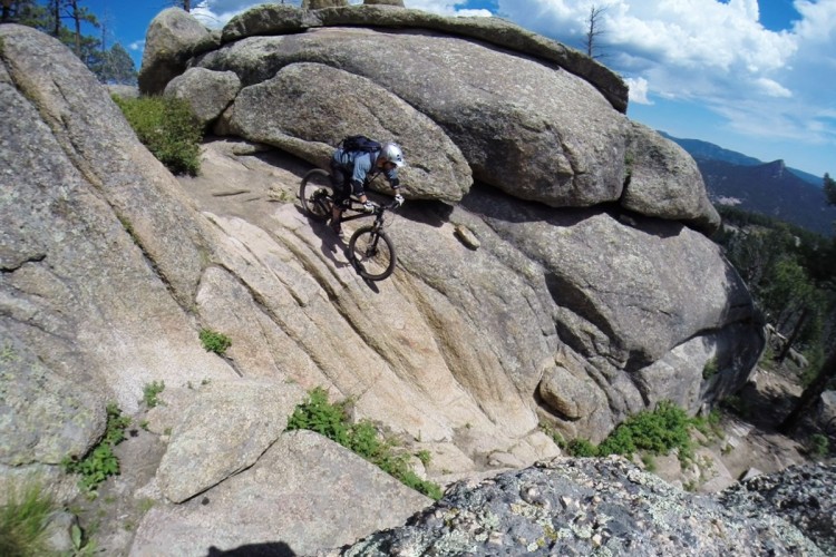 Mountain biker navigating a rocky and steep terrain with large boulders under a blue sky and scattered clouds. Lush green vegetation surrounds the rocky path, showcasing a natural outdoor adventure setting.