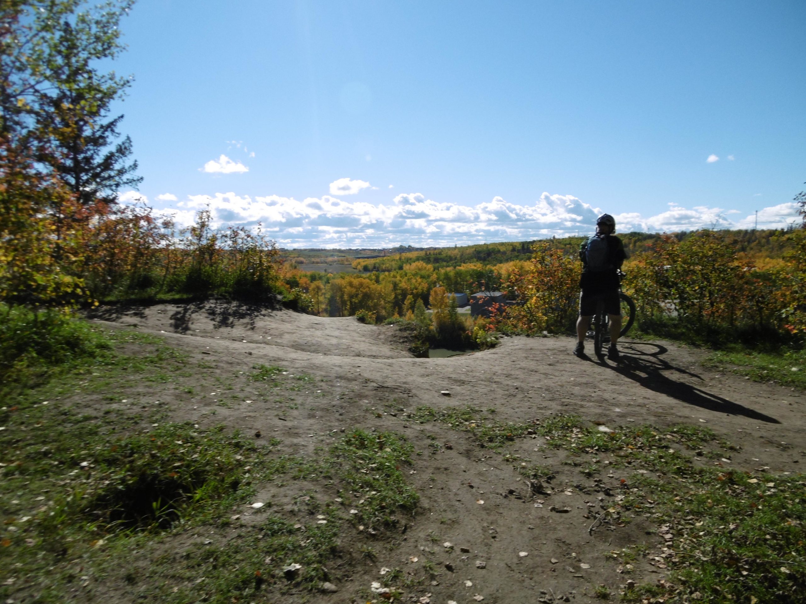 A person on a mountain bike stands at a scenic overlook, surrounded by autumn foliage. The landscape features rolling hills and a clear blue sky, with fluffy white clouds. The sun casts shadows on the dirt path, highlighting the natural beauty of the outdoor setting. Terwillegar Park mountain bike trail.