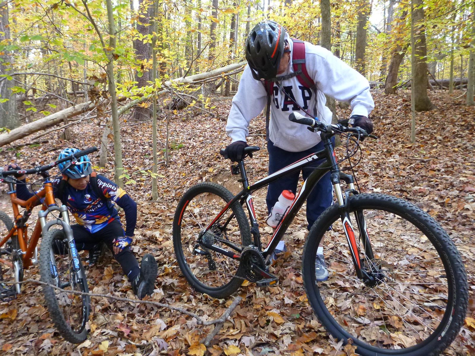 Two cyclists are positioned in a wooded area covered with fallen leaves. One cyclist is kneeling beside an orange mountain bike, appearing to adjust something, while the other cyclist stands nearby, focused on a black mountain bike. The setting features autumn foliage, with trees in the background, creating a natural environment for outdoor biking activities. Wild Turkey mountain bike trail.
