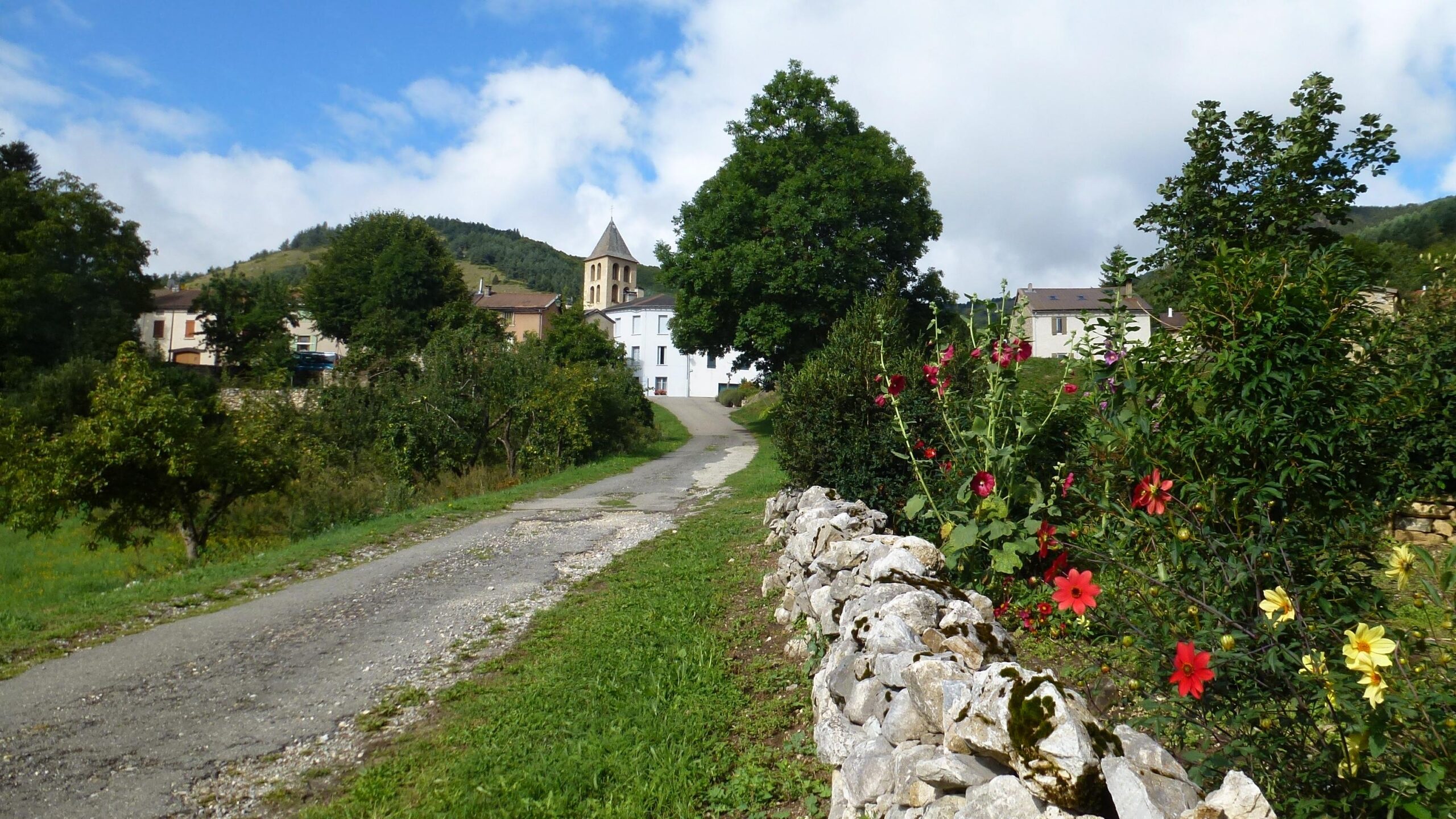 A picturesque rural scene featuring a winding gravel road leading towards a small village. On one side, a stone wall is lined with vibrant flowers in various colors. In the background, there are quaint buildings with a church tower visible, surrounded by lush greenery and rolling hills under a partly cloudy sky. Great Crossing of the Ariege mountain bike trail.