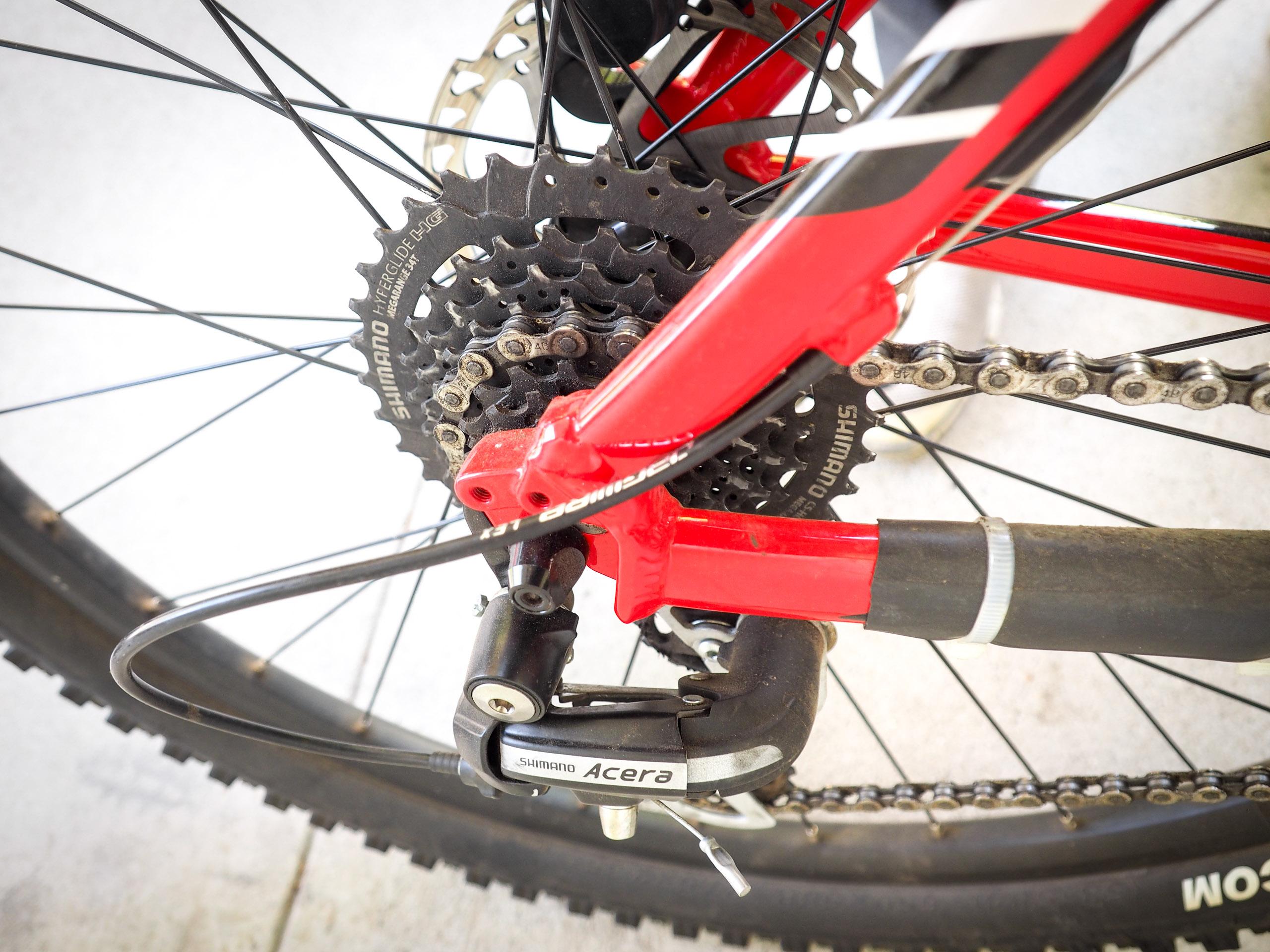 Giant Talon 27.5 4: Close-up view of a red mountain bike's rear drivetrain, featuring a Shimano Acera derailleur, chain, and cassette. The background is a light surface, and the bike's wheel is partially visible.