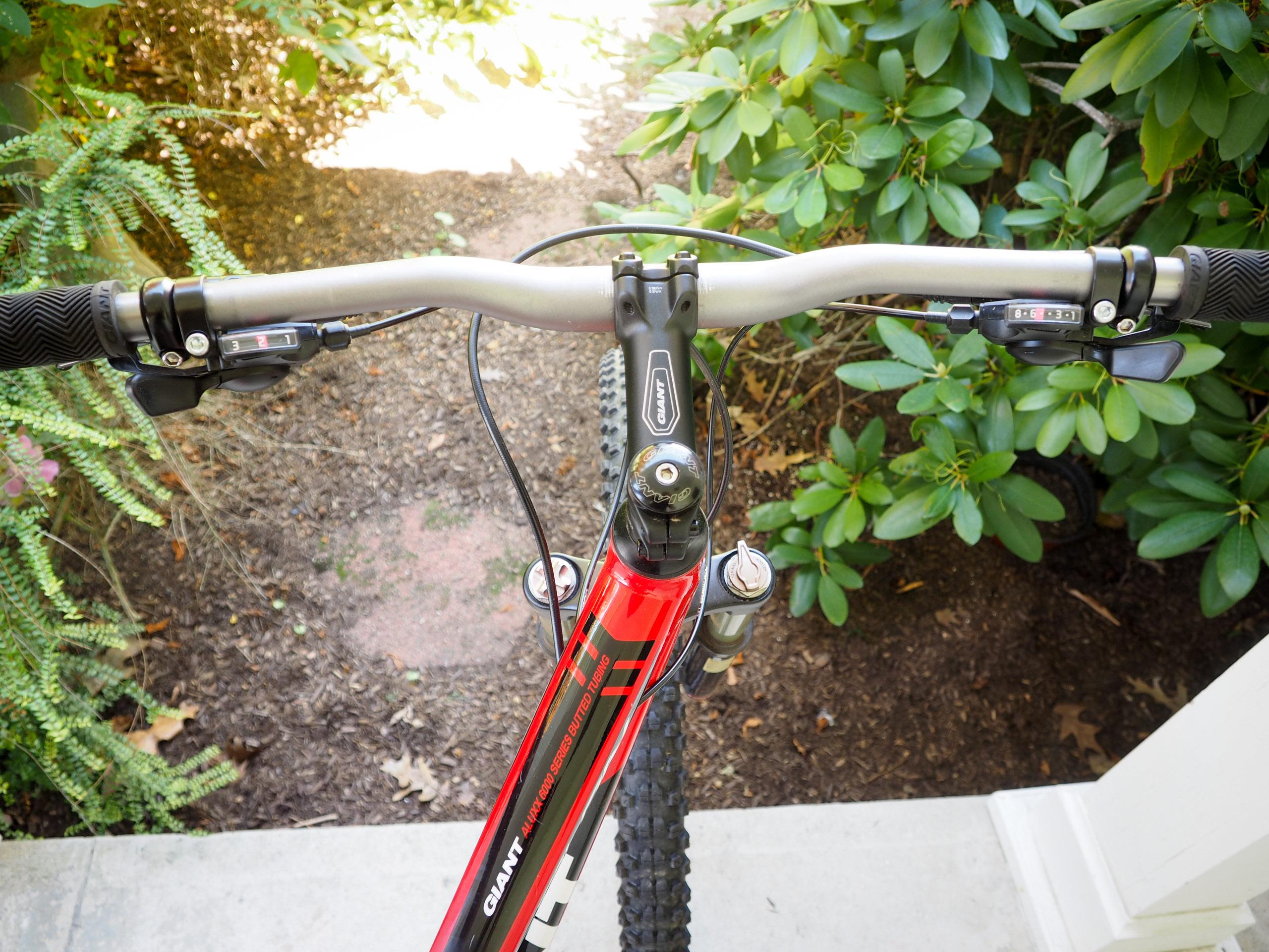 Giant Talon 27.5 4: Alt text: A close-up view of a mountain bike's handlebars, featuring gear shifters and brake levers, set against a backdrop of greenery and gravel. The bike has a red frame and the brand name "Giant" is visible on the handlebars.