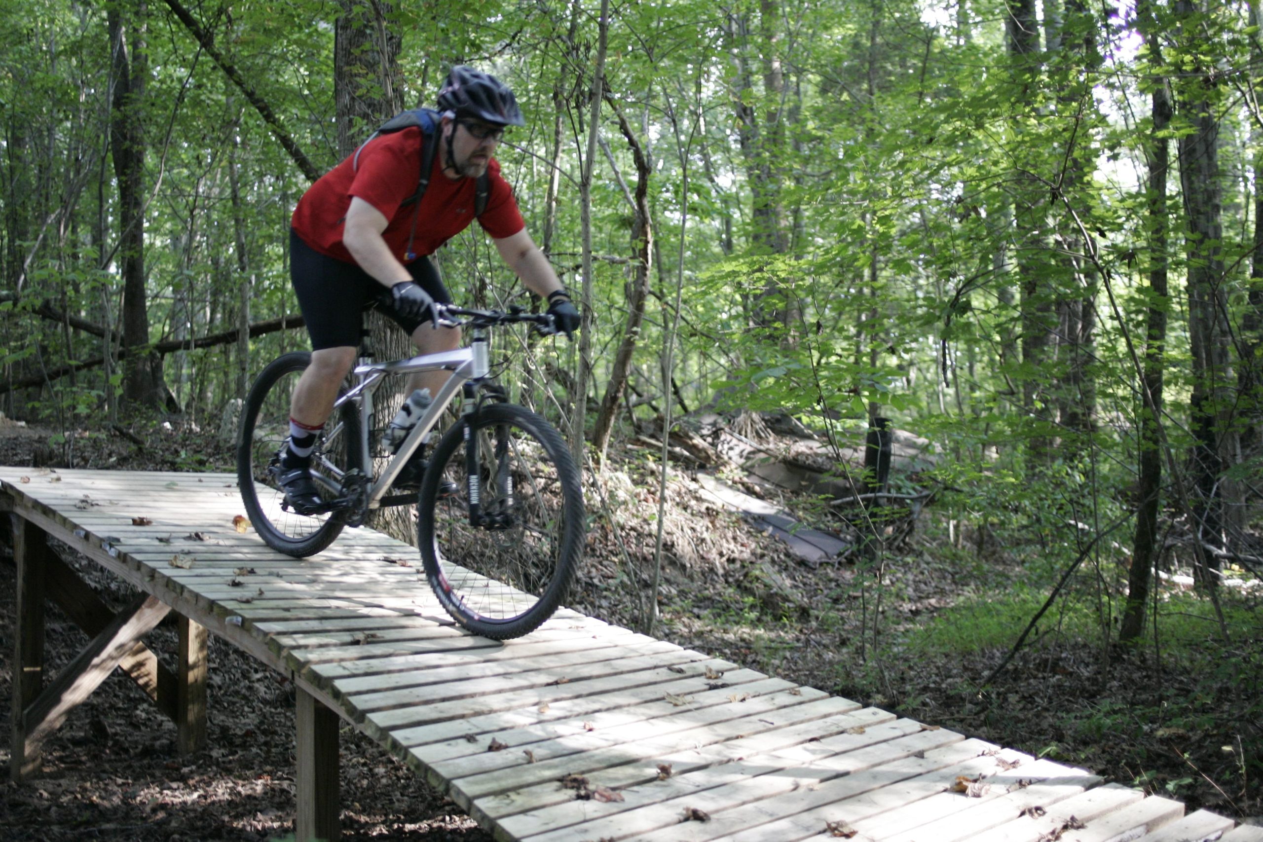 A mountain biker in a red shirt rides along a wooden bridge in a forested area, surrounded by green trees and foliage. Kernersville MTB park mountain bike trail.