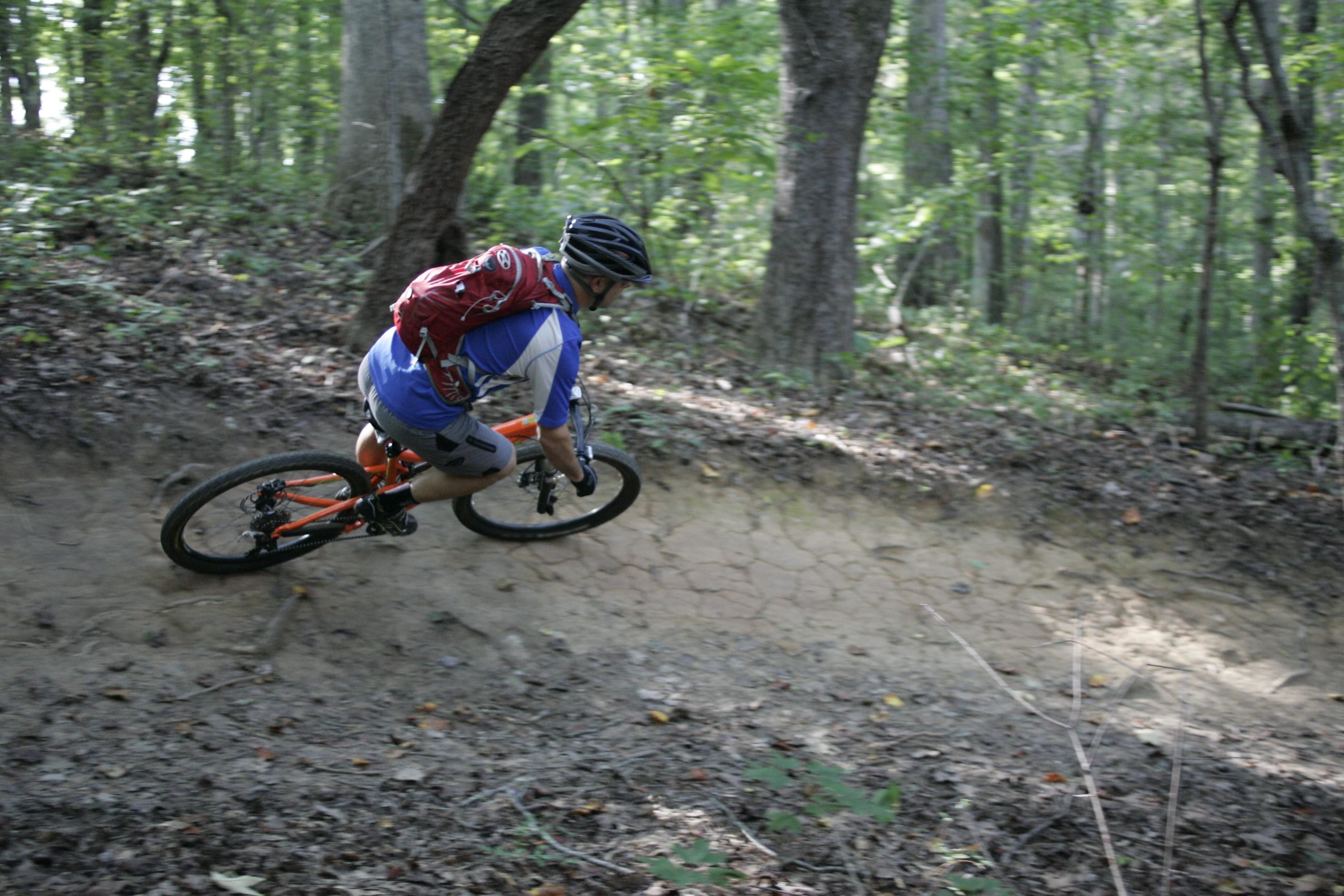 A mountain biker leans into a turn while navigating a dirt trail surrounded by trees and foliage. The biker wears a helmet and a backpack, showcasing an intense focus on the trail. Sunlight filters through the forest, highlighting the rugged terrain. Kernersville MTB park mountain bike trail.