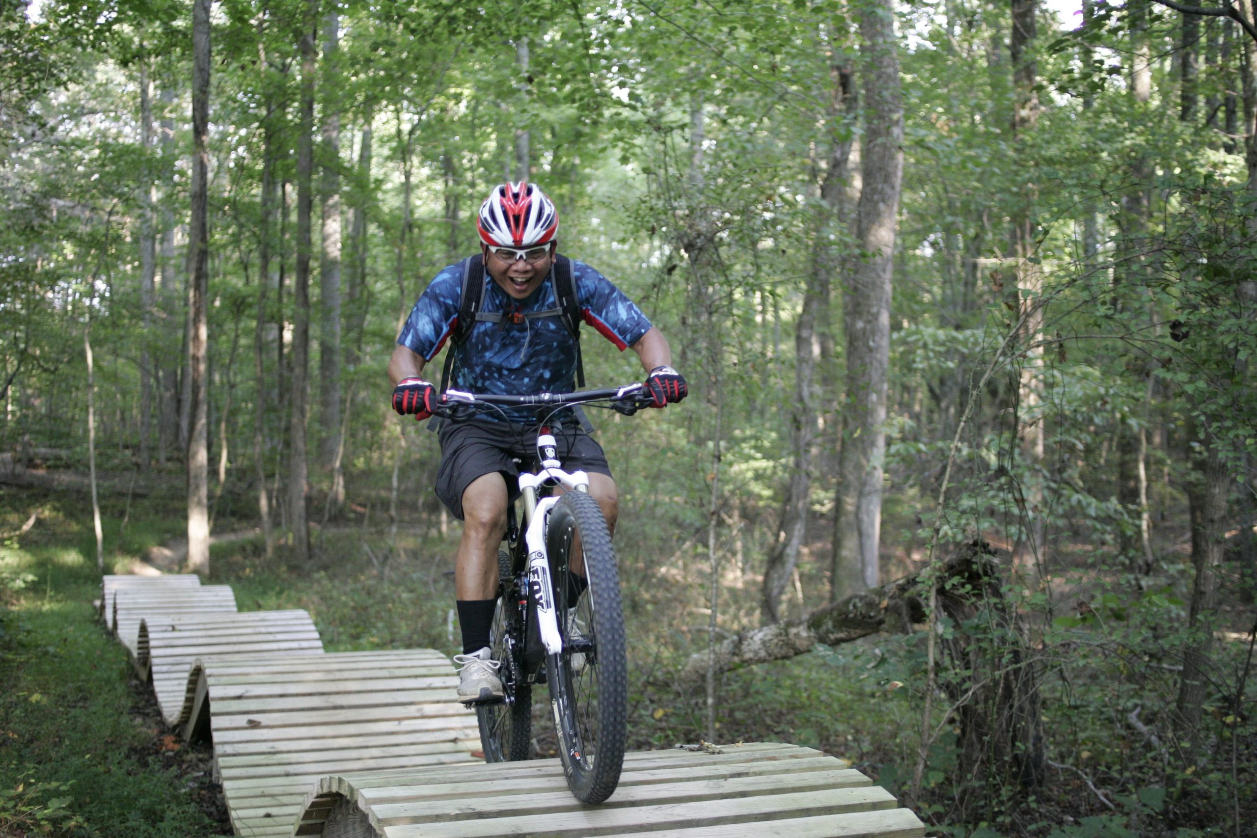 A mountain biker joyfully riding on a wooden trail path through a lush green forest. The cyclist is wearing a helmet and colorful gear, capturing the excitement of the ride. Surrounding trees and foliage create a vibrant natural backdrop. Kernersville MTB park mountain bike trail.