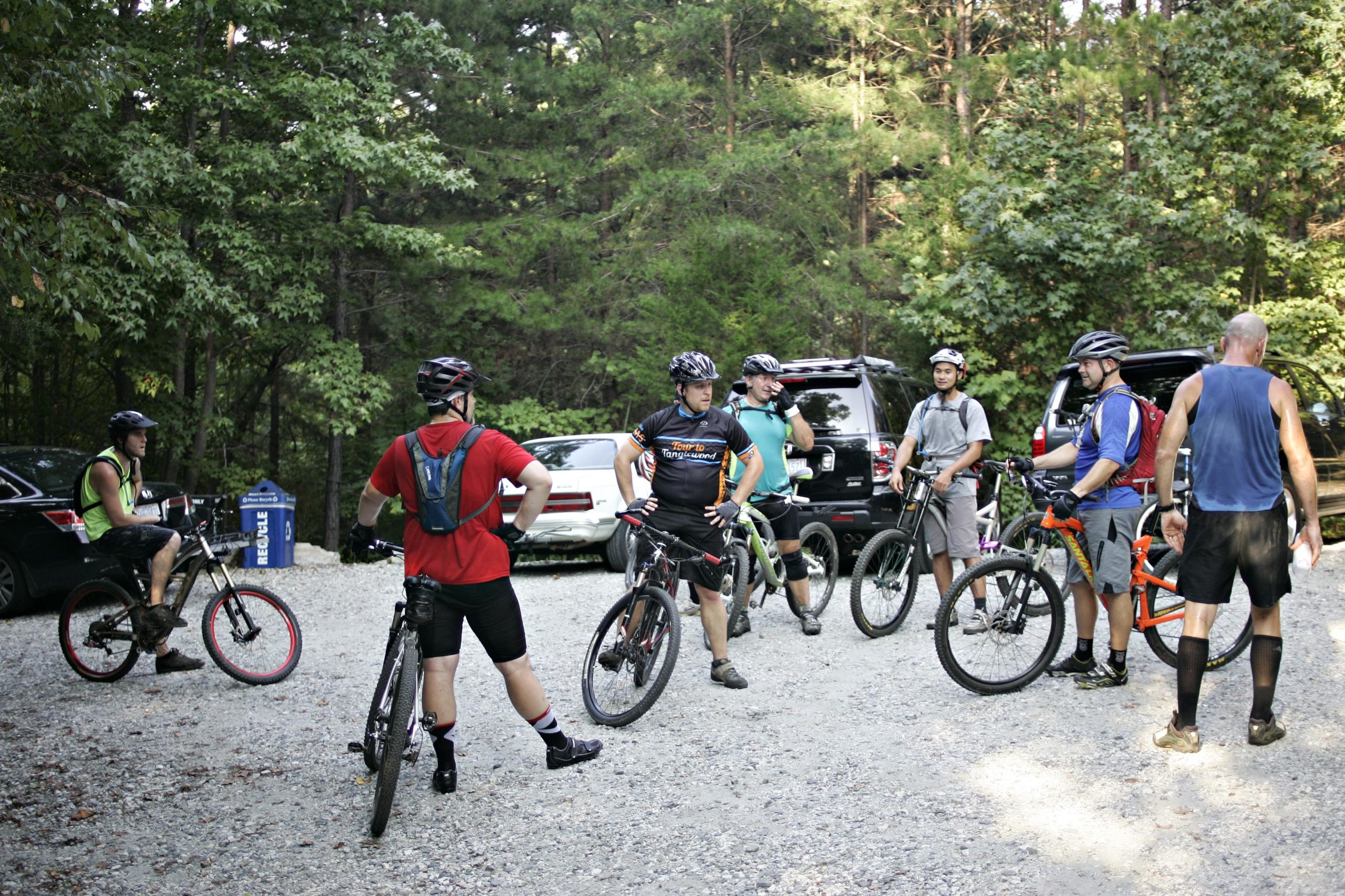 A group of six mountain bikers in a gravel parking area surrounded by trees, preparing for a ride. They are wearing helmets and cycling gear, with their bikes nearby. Some are engaged in conversation, while others appear to be adjusting their gear. In the background, parked vehicles are visible. Kernersville MTB park mountain bike trail.