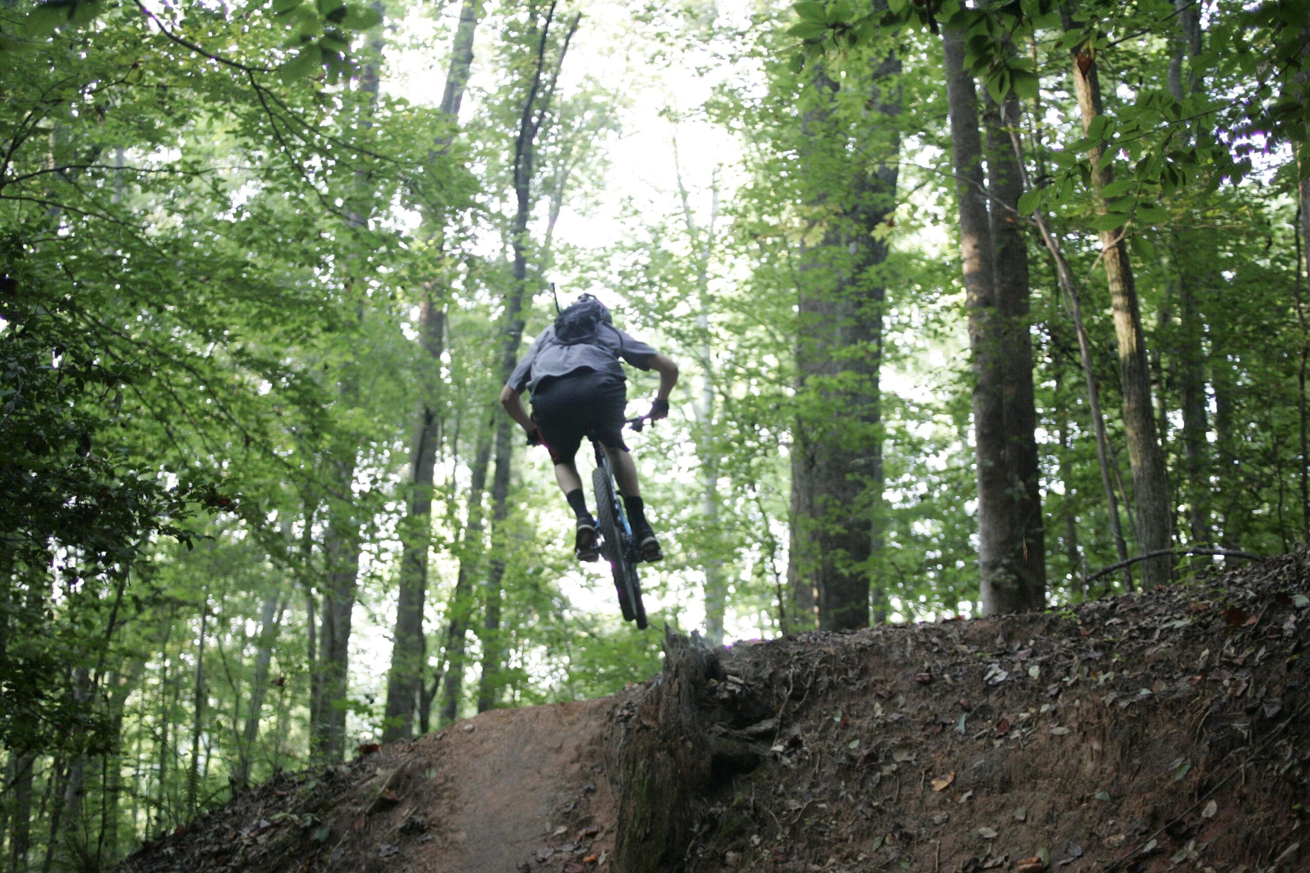 A mountain biker performs a jump off a dirt ramp in a forested area, surrounded by green trees and foliage. The rider is in the air, showcasing a dynamic pose as they navigate the terrain. Salem Lake mountain bike trail.