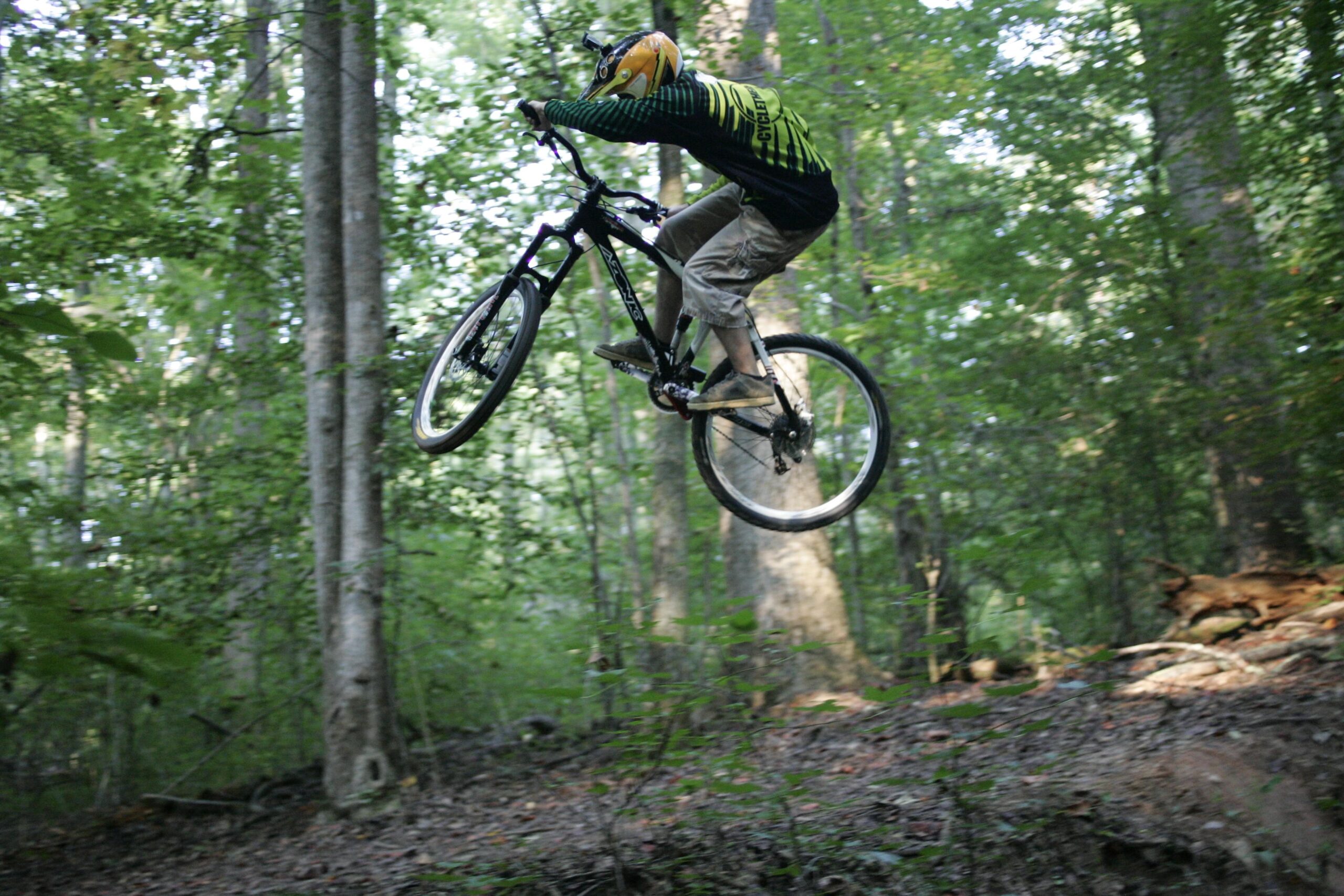 A person riding a mountain bike is mid-air while performing a jump on a wooded trail, surrounded by tall trees and lush greenery. The cyclist is wearing a helmet and a jersey with a bold design, showcasing an adventurous moment in outdoor biking. Salem Lake mountain bike trail.