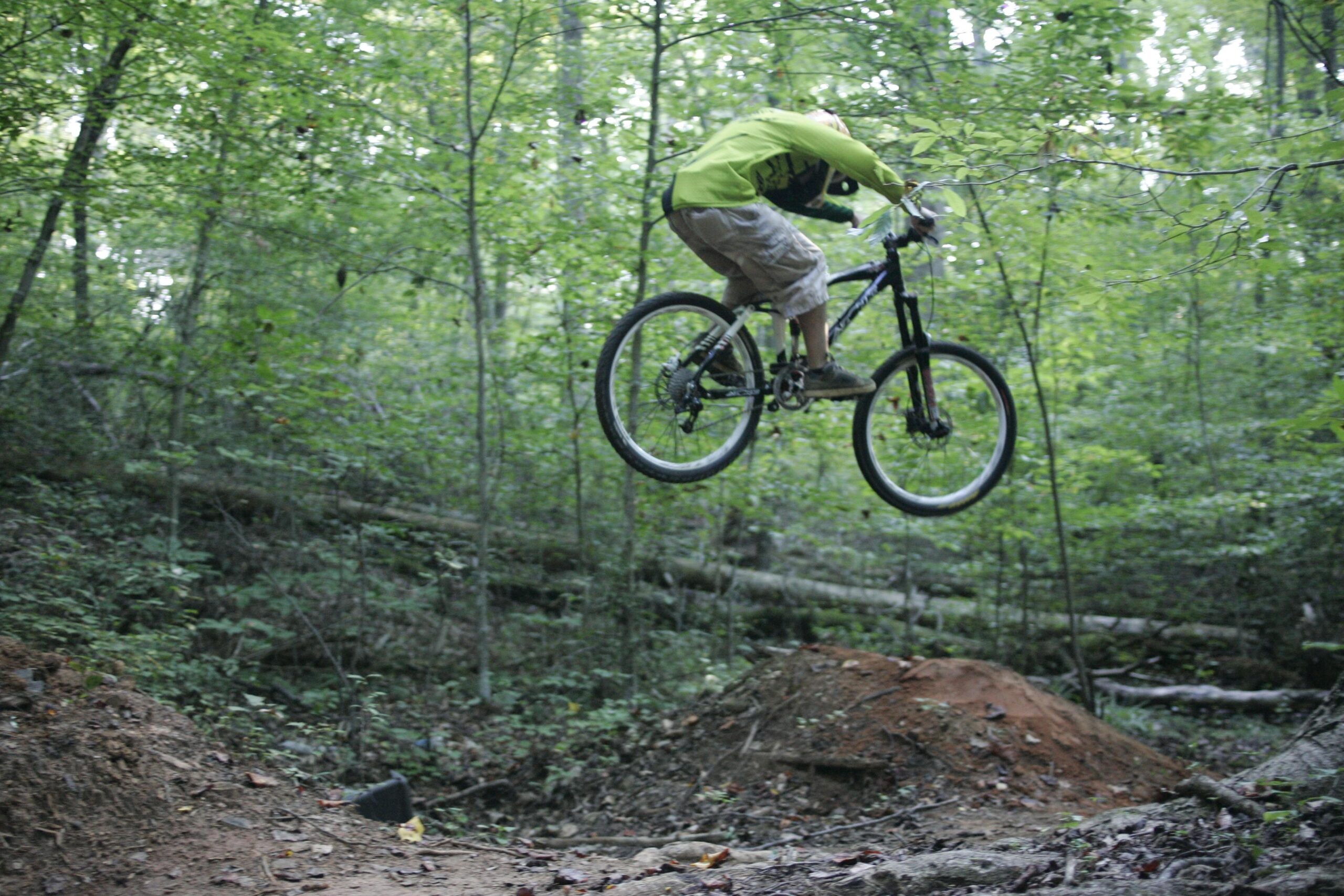 A person in a bright green shirt riding a mountain bike jumps off a dirt ramp in a wooded area. The surrounding forest is lush with greenery, and fallen logs can be seen in the background. Salem Lake mountain bike trail.