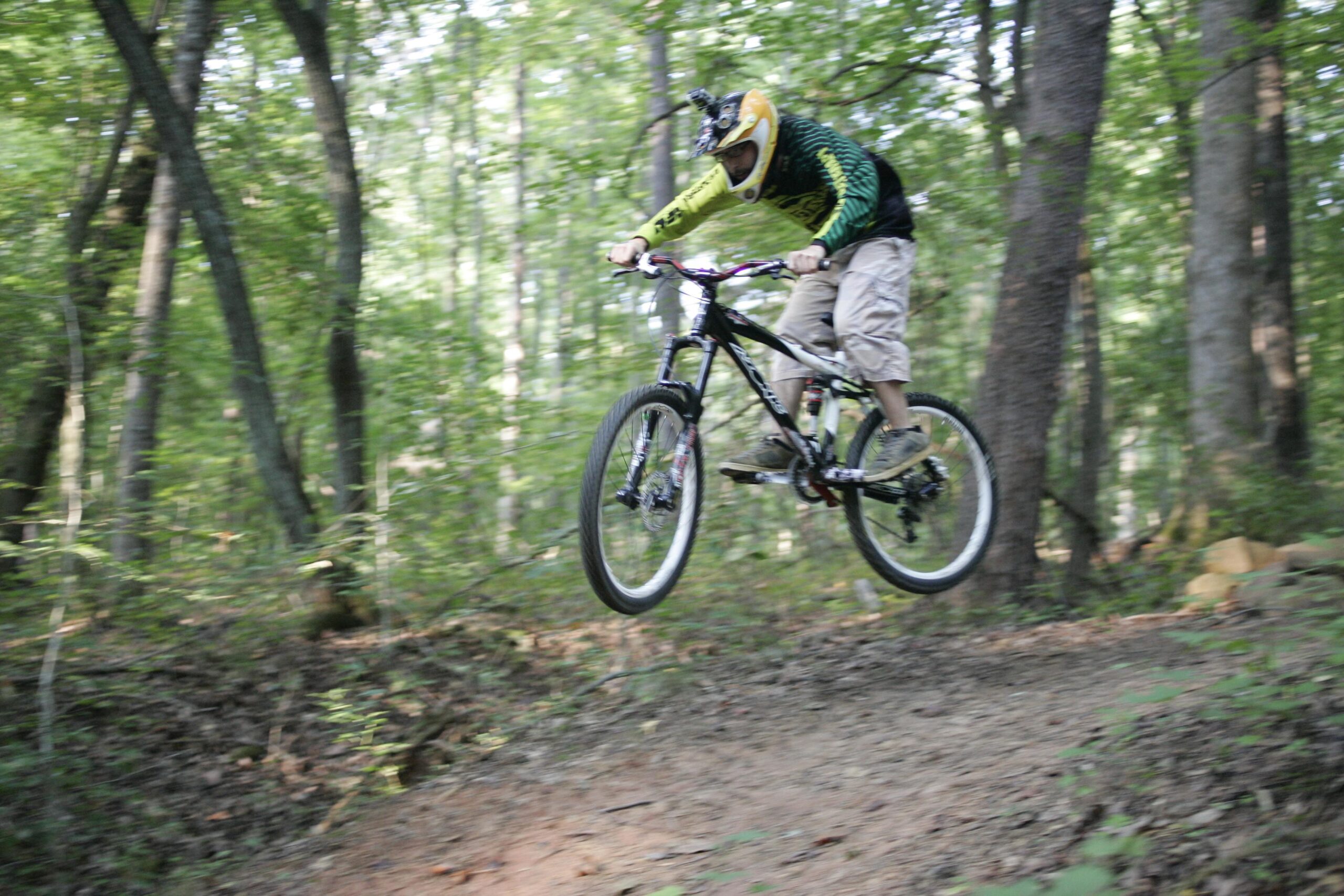 A mountain biker wearing a helmet and protective gear performs a jump on a trail in a lush, wooded area. The bike is airborne, with a focus on the rider's posture and technique as they navigate the trail. Salem Lake mountain bike trail.