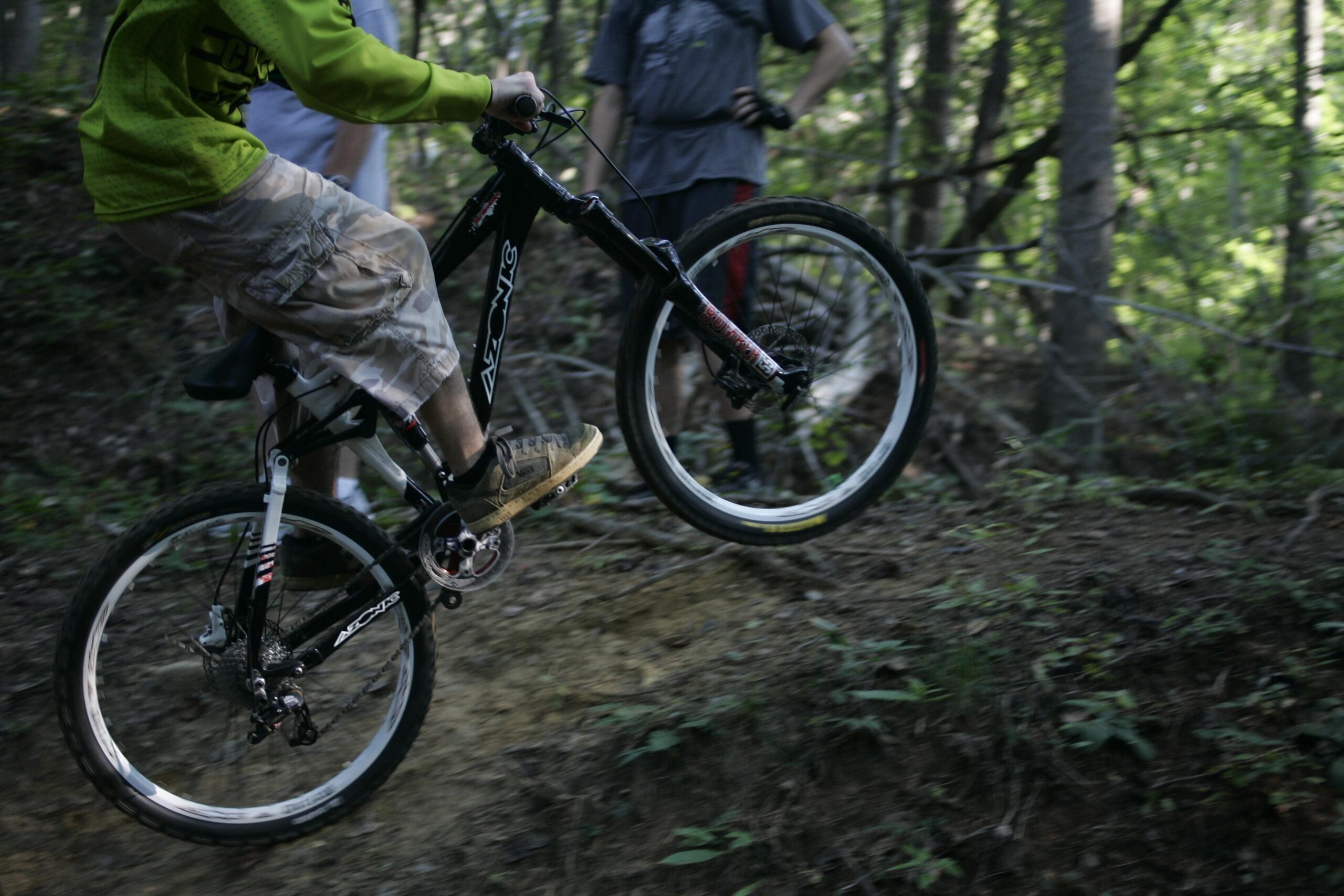 A person in a green shirt is performing a jump on a mountain bike, with the front wheel lifted off the ground. In the background, two other individuals watch in a forested area. The terrain is uneven and covered with dirt and greenery. Salem Lake mountain bike trail.