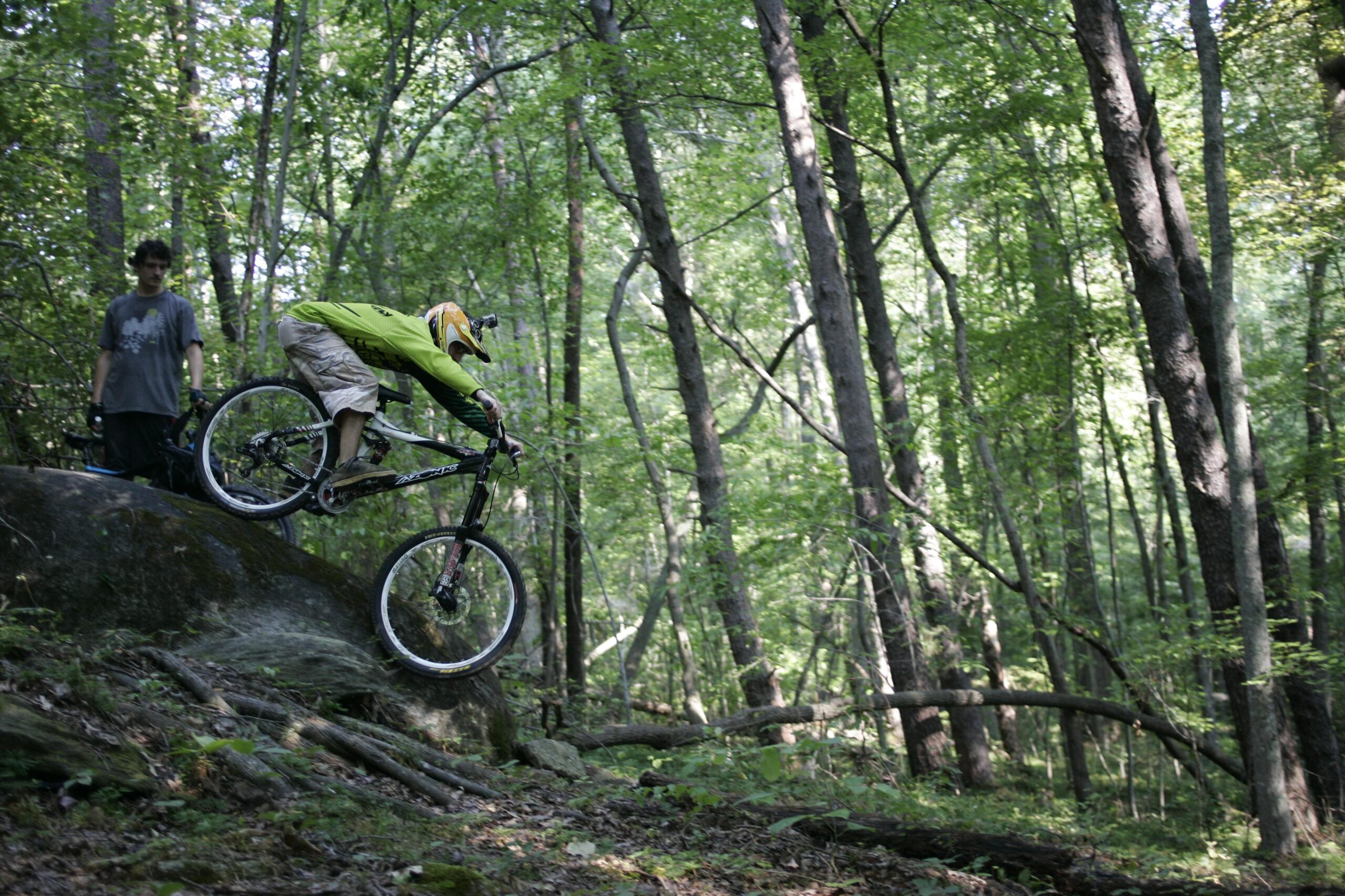 A mountain biker in a bright green jacket and helmet jumps off a large rock in a lush green forest, while a second person watches from behind. The scene captures the thrill of mountain biking in a natural setting. Salem Lake mountain bike trail.