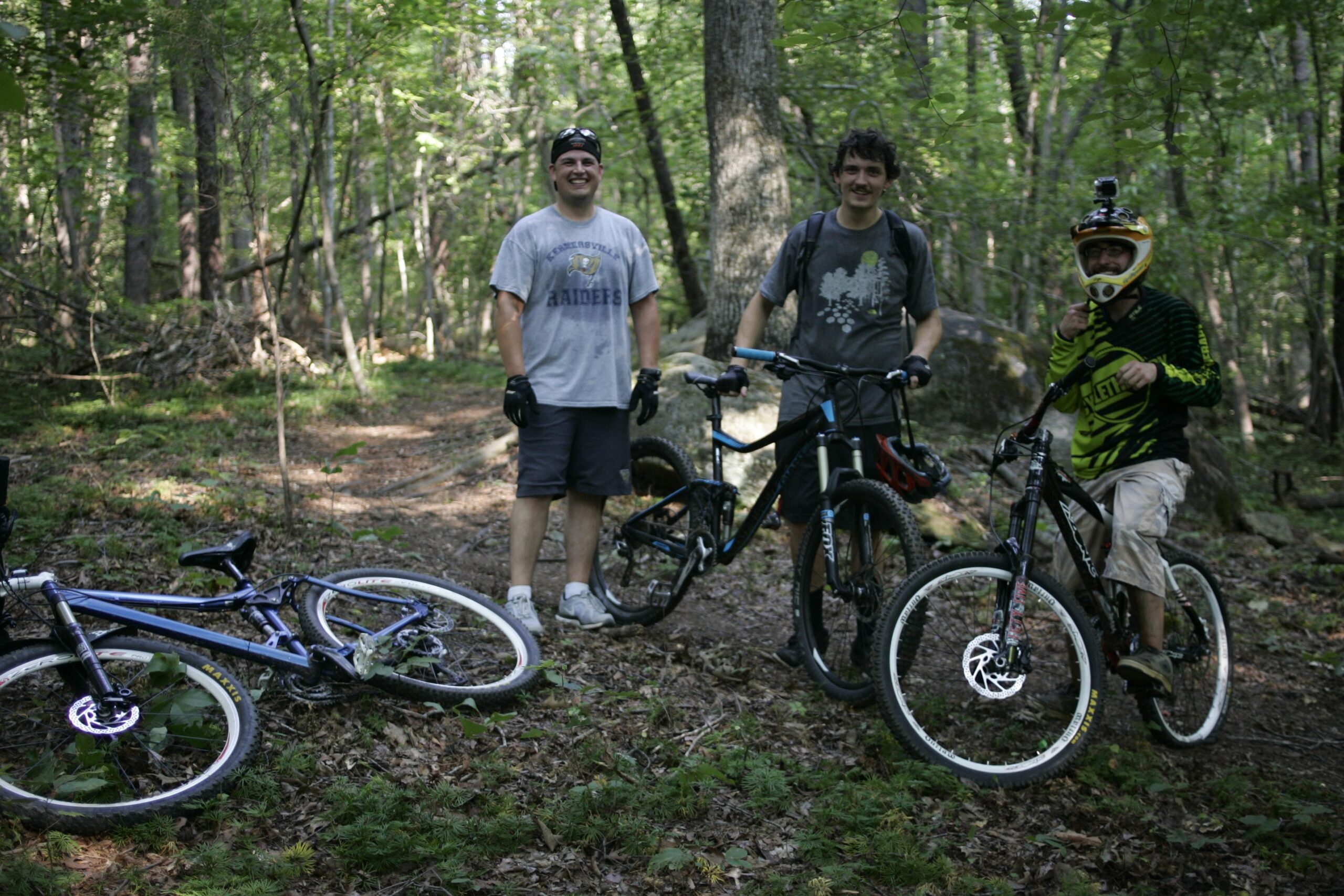 Three mountain bikers posing in a forested area, surrounded by trees and greenery. Two riders stand next to their bikes, while another is seated on a bike with a helmet. One bike is on the ground nearby. The scene captures a sense of adventure and camaraderie among the cyclists. Salem Lake mountain bike trail.