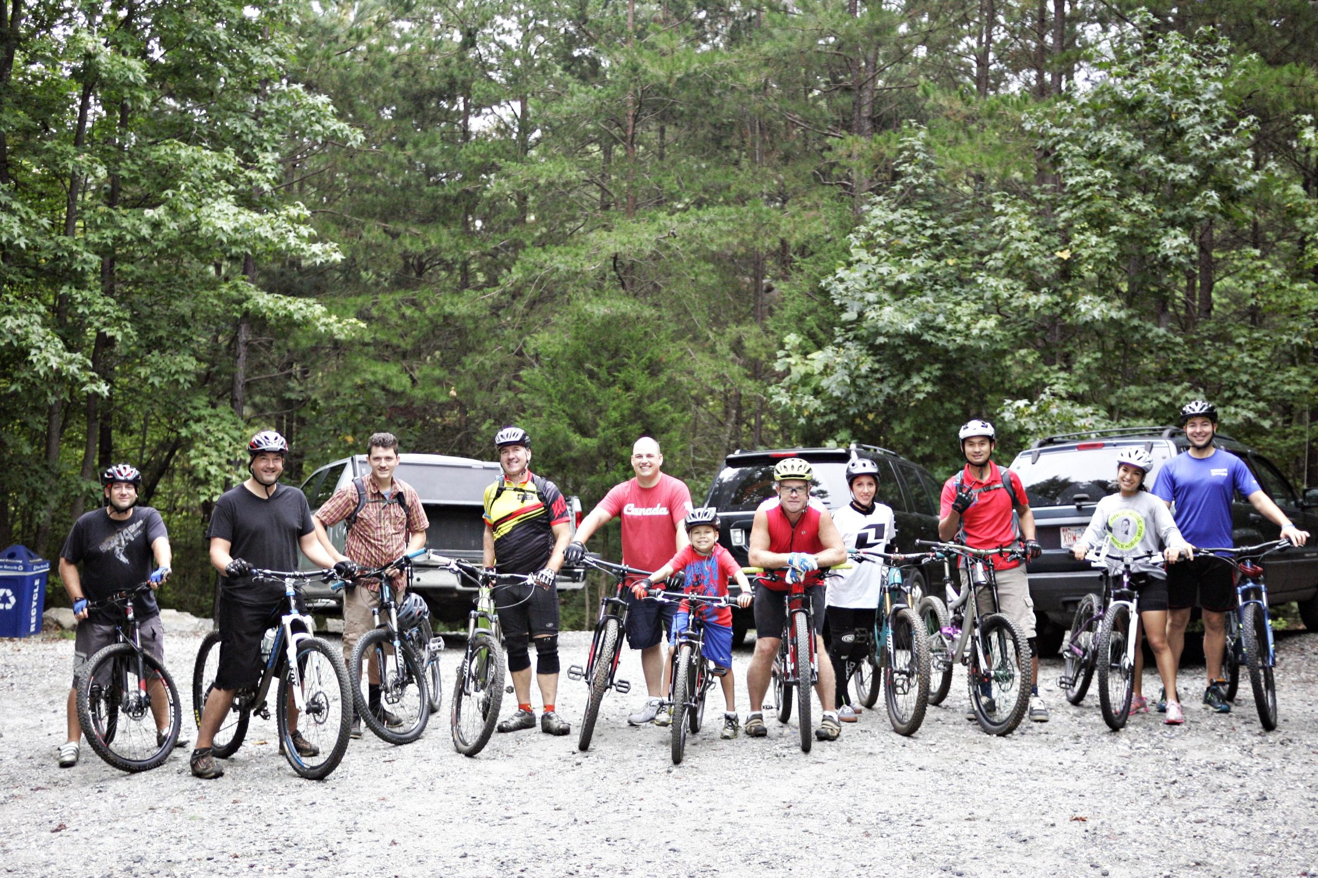 A group of ten mountain bikers stands on a gravel path surrounded by lush trees, each person holding a bike. They are wearing helmets and a variety of colorful cycling gear. In the background, there are parked cars hinting at a biking excursion. The mood is cheerful and adventurous. Kernersville MTB park mountain bike trail.