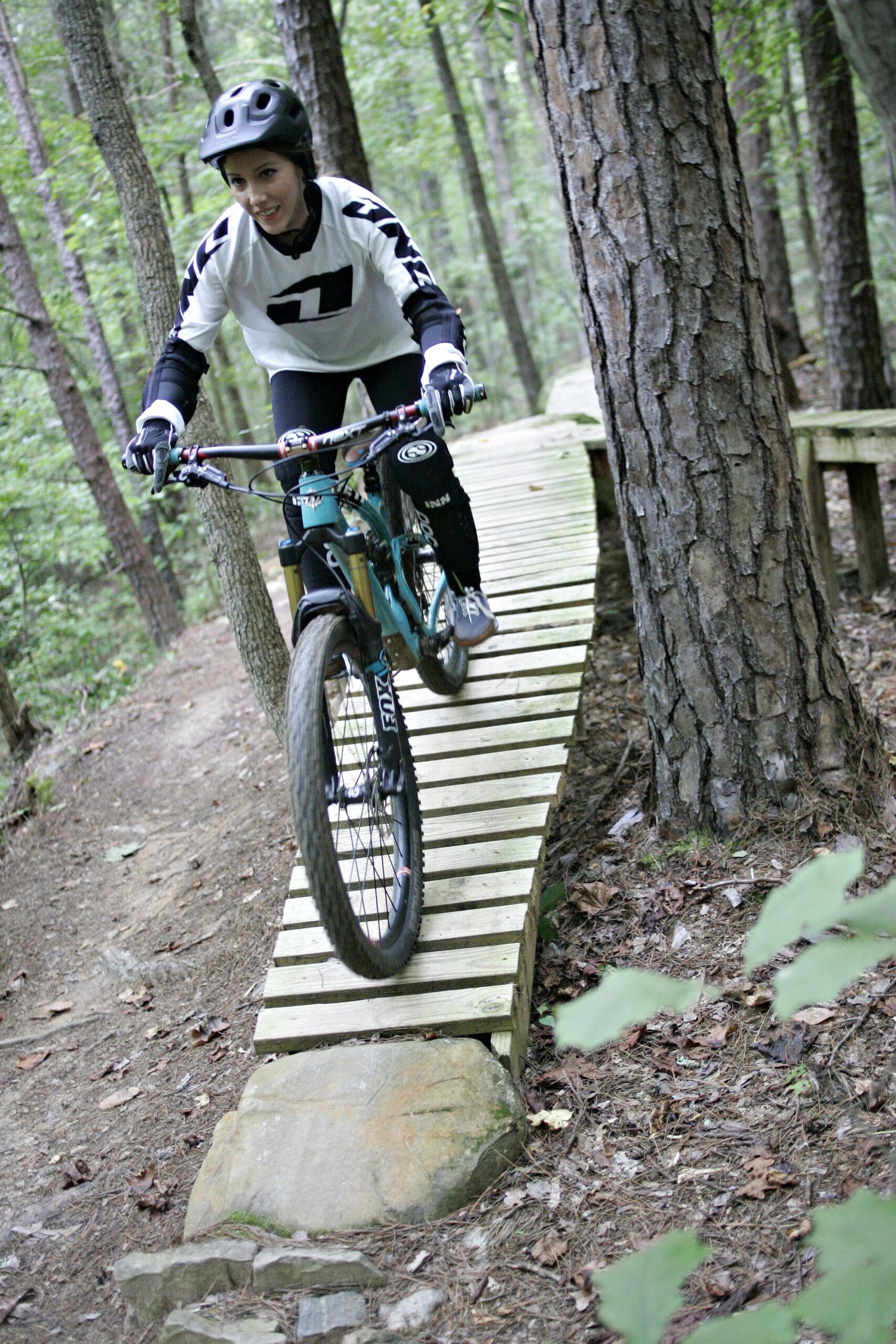 A mountain biker navigating a wooden bridge along a trail in a wooded area, wearing a helmet and protective gear. The scene captures the excitement of biking through nature, with a focus on the rider's skill and the lush greenery surrounding the path. Kernersville MTB park mountain bike trail.