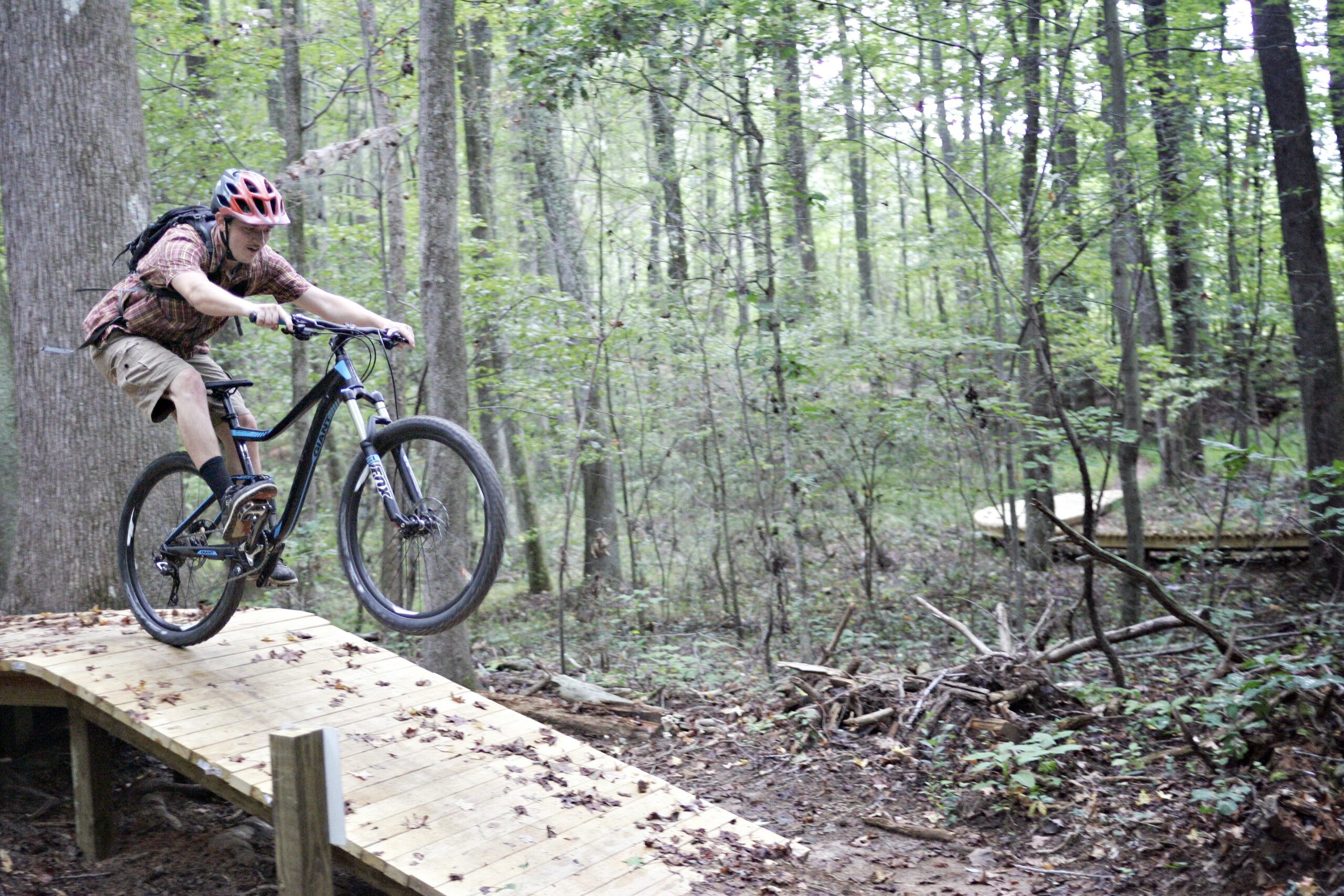 A mountain biker in a plaid shirt and helmet performs a jump on a wooden trail in a forested area, surrounded by trees and foliage. Kernersville MTB park mountain bike trail.