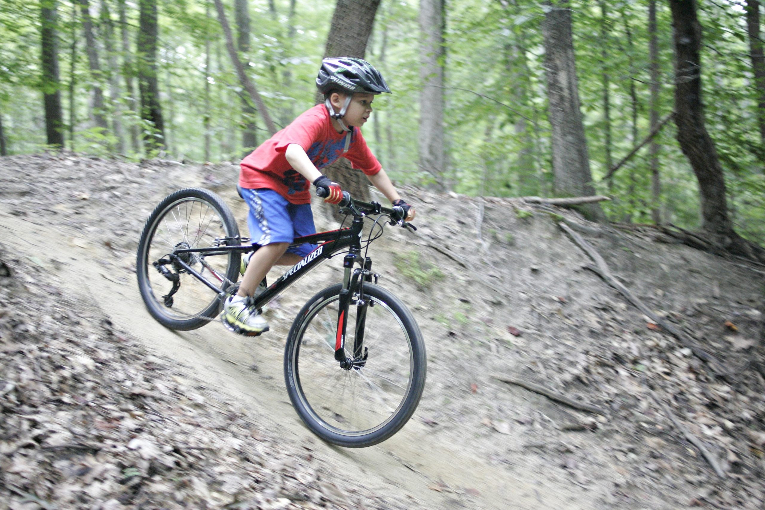 A young boy riding a mountain bike down a dirt trail in a wooded area, wearing a helmet and a red t-shirt with blue shorts. He appears to be focused and energized as he navigates the incline. Surrounding foliage includes green trees and fallen leaves on the ground. Kernersville MTB park mountain bike trail.