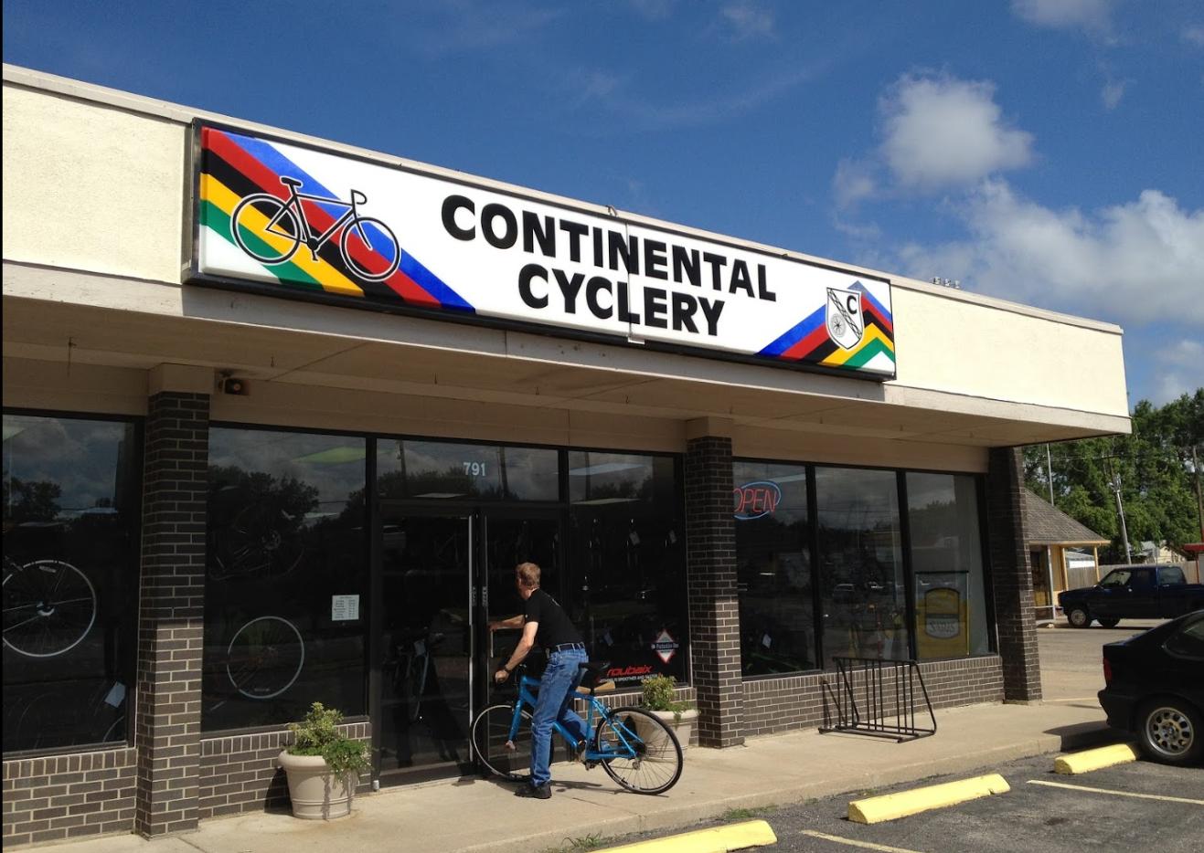 A bicycle shop storefront labeled "Continental Cyclery" with a colorful bike logo above the entrance. A person is seen entering the shop while riding a blue bicycle, and the shop windows display various bicycles. The background includes a clear blue sky and nearby vehicles parked outside.