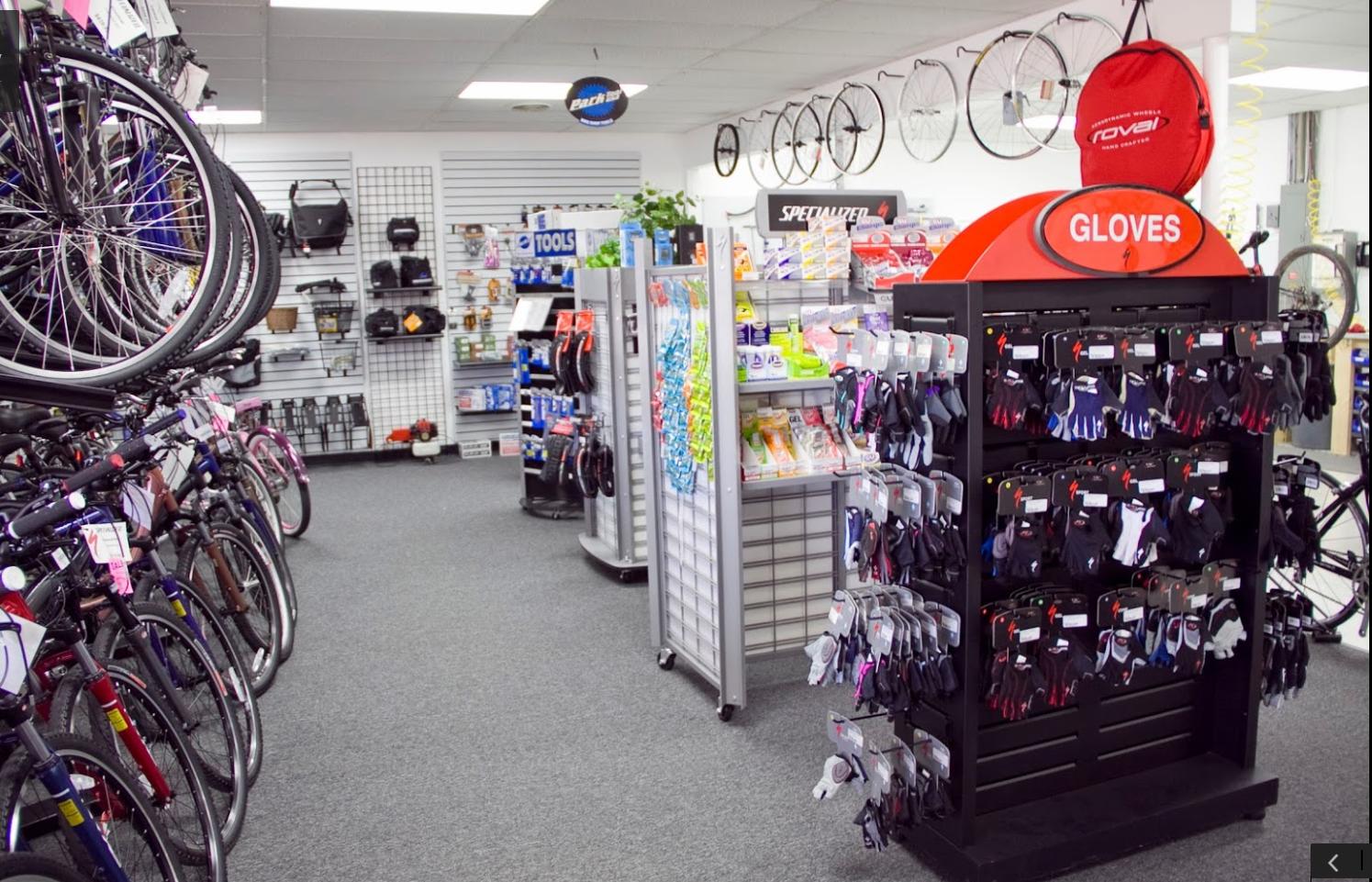 A view inside a bicycle shop featuring a variety of bikes displayed along the walls and shelves stocked with cycling accessories. On the right, a prominent display rack showcases different styles of cycling gloves. The shop has a clean, organized layout with gray carpet flooring and walls adorned with bicycle wheels and various cycling gear.