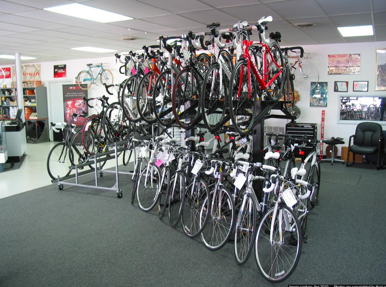 A well-organized bicycle shop featuring a variety of bicycles on display. The upper level showcases several racing bikes in different colors, while the lower level has a selection of road bikes. The shop has a clean, spacious layout with a gray carpet and bright lighting, enhancing the visibility of the bikes and accessories.