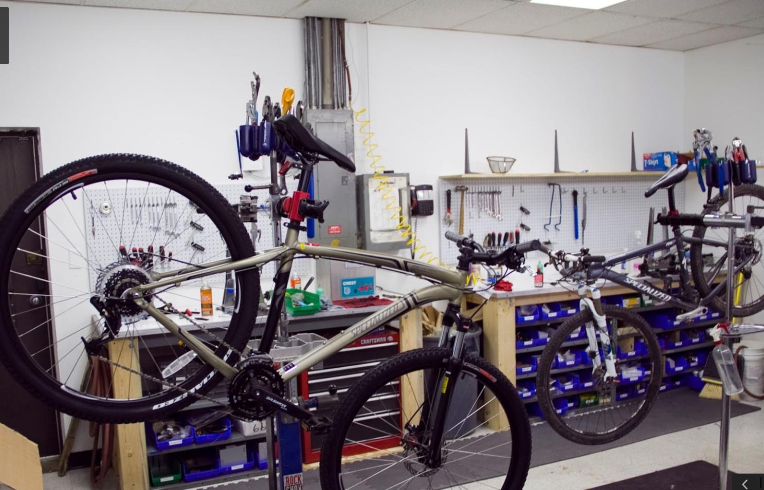 A bike repair workshop featuring two bicycles on repair stands, with one bike upside down and the other upright. The background includes a pegboard with various tools and bike parts, as well as storage bins. The space is well-organized and equipped for bicycle maintenance.