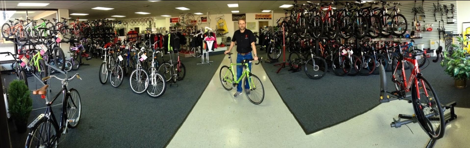A panoramic view of a bicycle shop interior, showcasing a wide selection of bicycles and cycling gear. The space is filled with various bikes, including colorful options displayed on racks and stands. A man stands in the foreground holding a bright green bicycle, surrounded by additional bikes and cycling apparel hanging on display. The flooring is carpeted, and the shop has a bright, well-lit atmosphere.
