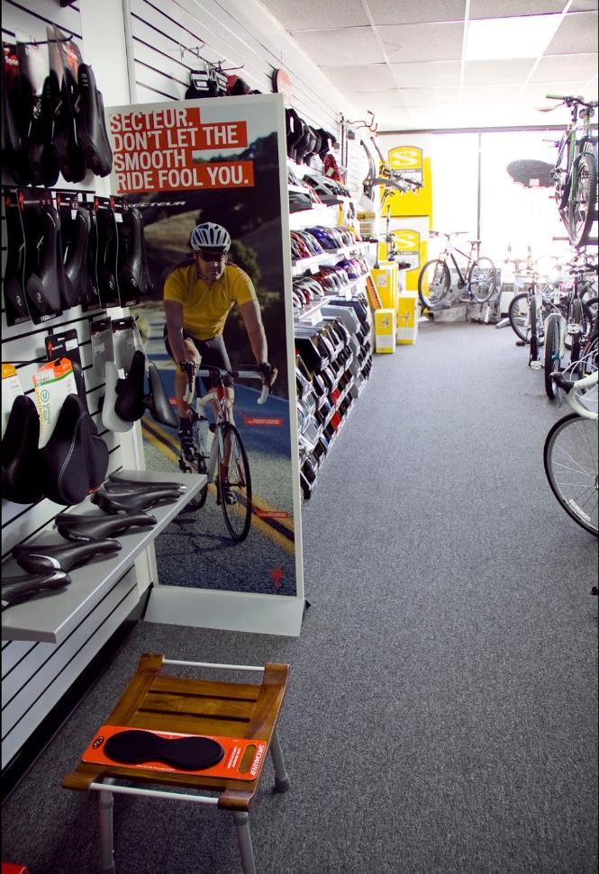 A view of a bicycle shop interior featuring a variety of bicycle seats displayed on the wall. In the foreground, there is a wooden stool with a bicycle seat accessory on it. A promotional banner stands next to the aisle, showcasing a cyclist with the text "Secteur. Don't let the smooth ride fool you." Bicycles and additional cycling gear can be seen in the background.